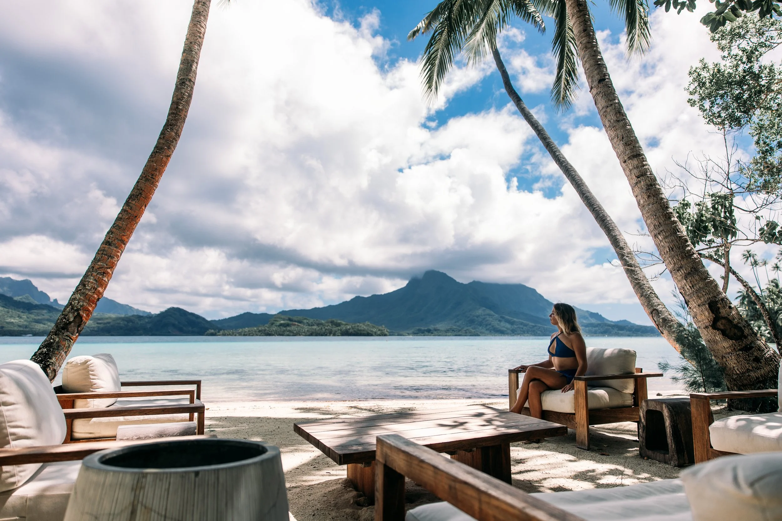 Une femme en maillot de bain assise sur un canapé en bois sur une plage de sable blanc, avec des palmiers inclinés de chaque côté et une montagne en arrière-plan, sous un ciel nuageux avec quelques zones de ciel bleu.