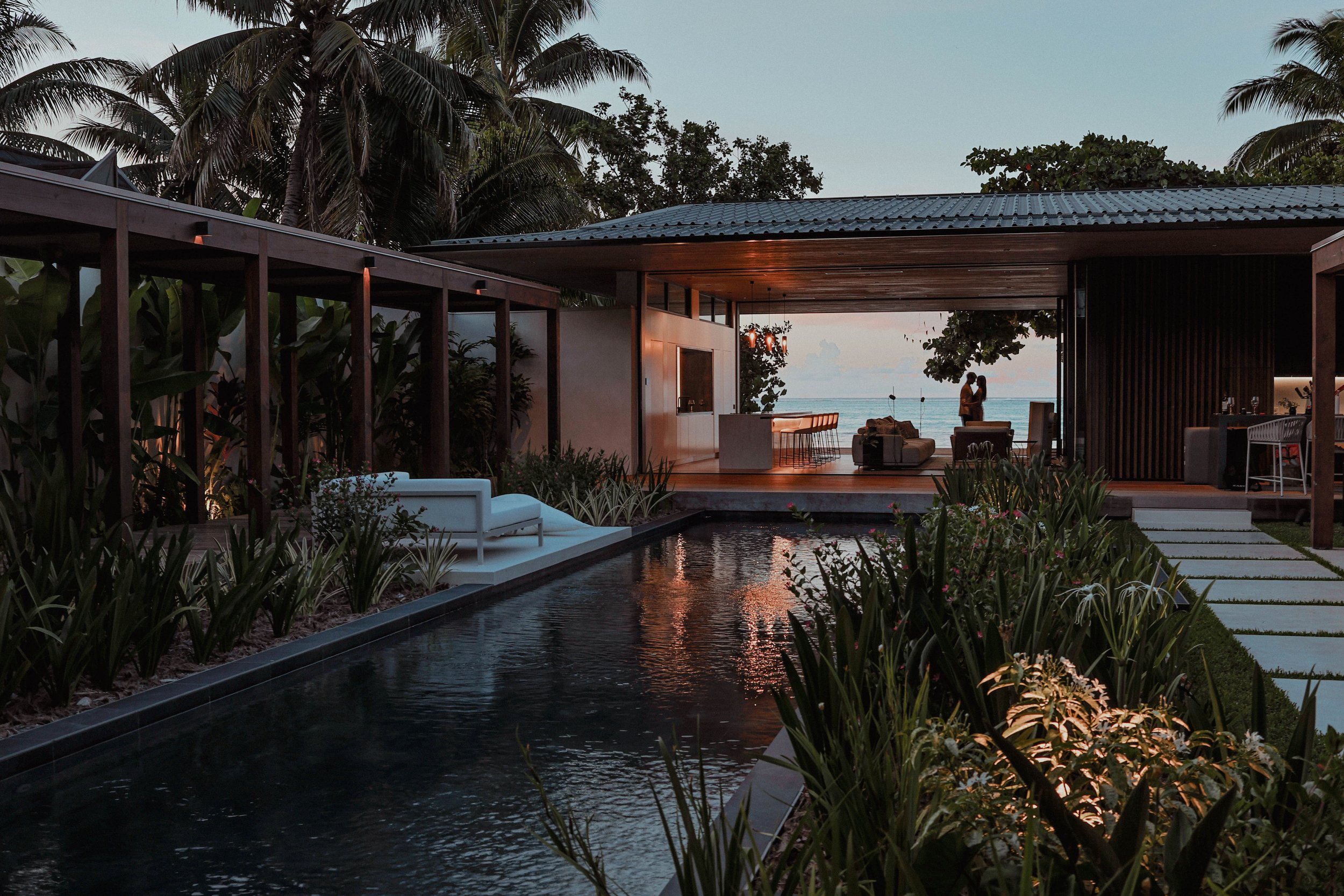 Une villa moderne en bord de mer avec une piscine étroite en avant-plan, entourée de végétation luxuriante et de palmiers. À l'arrière, un espace salon avec deux personnes qui semblent s'embrasser, face à l'océan au coucher du soleil.