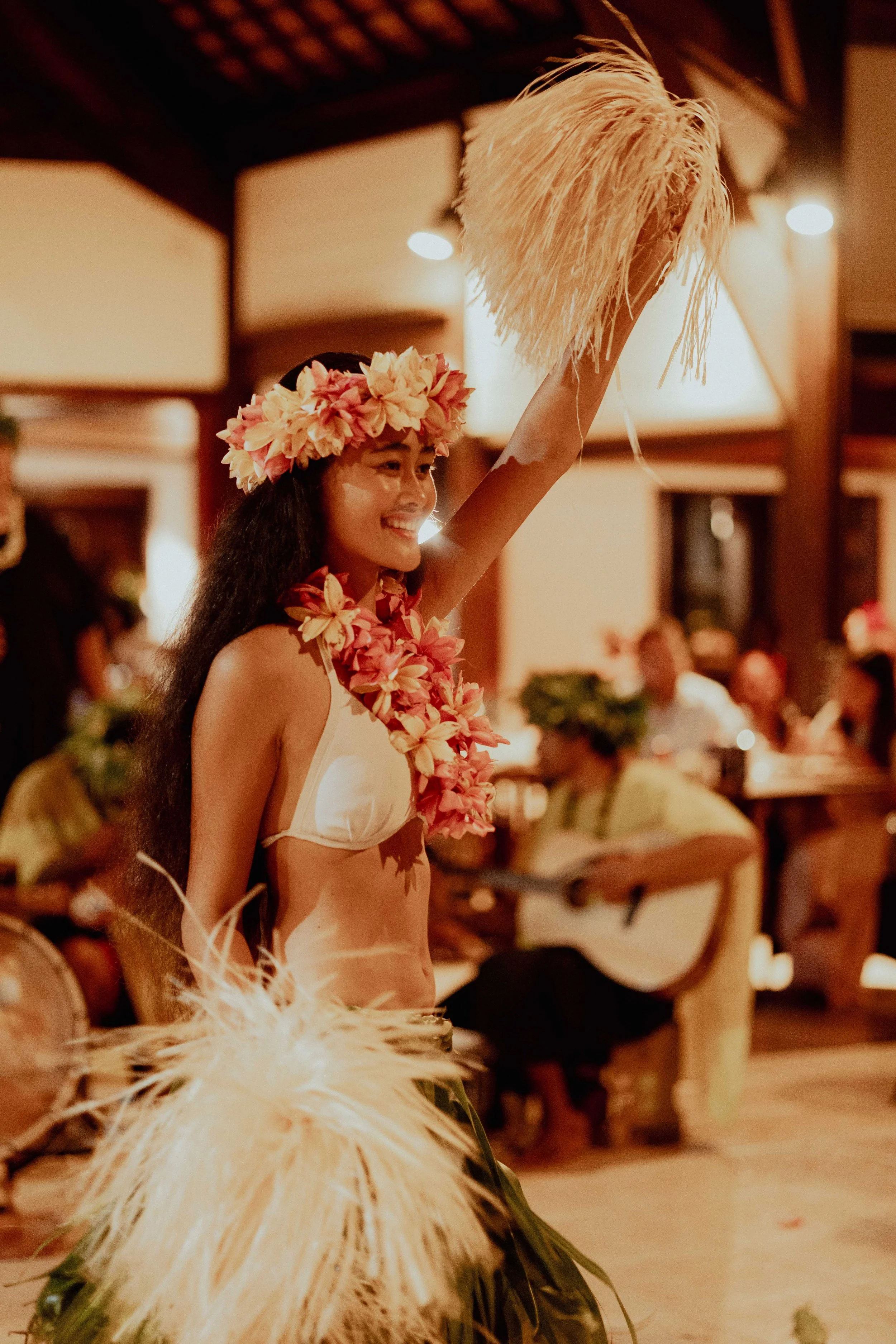 Jeune femme hawaïenne en costume traditionnel avec un collier de fleurs et une couronne de fleurs sur la tête, levant une large plume dans une ambiance de fête ou de danse