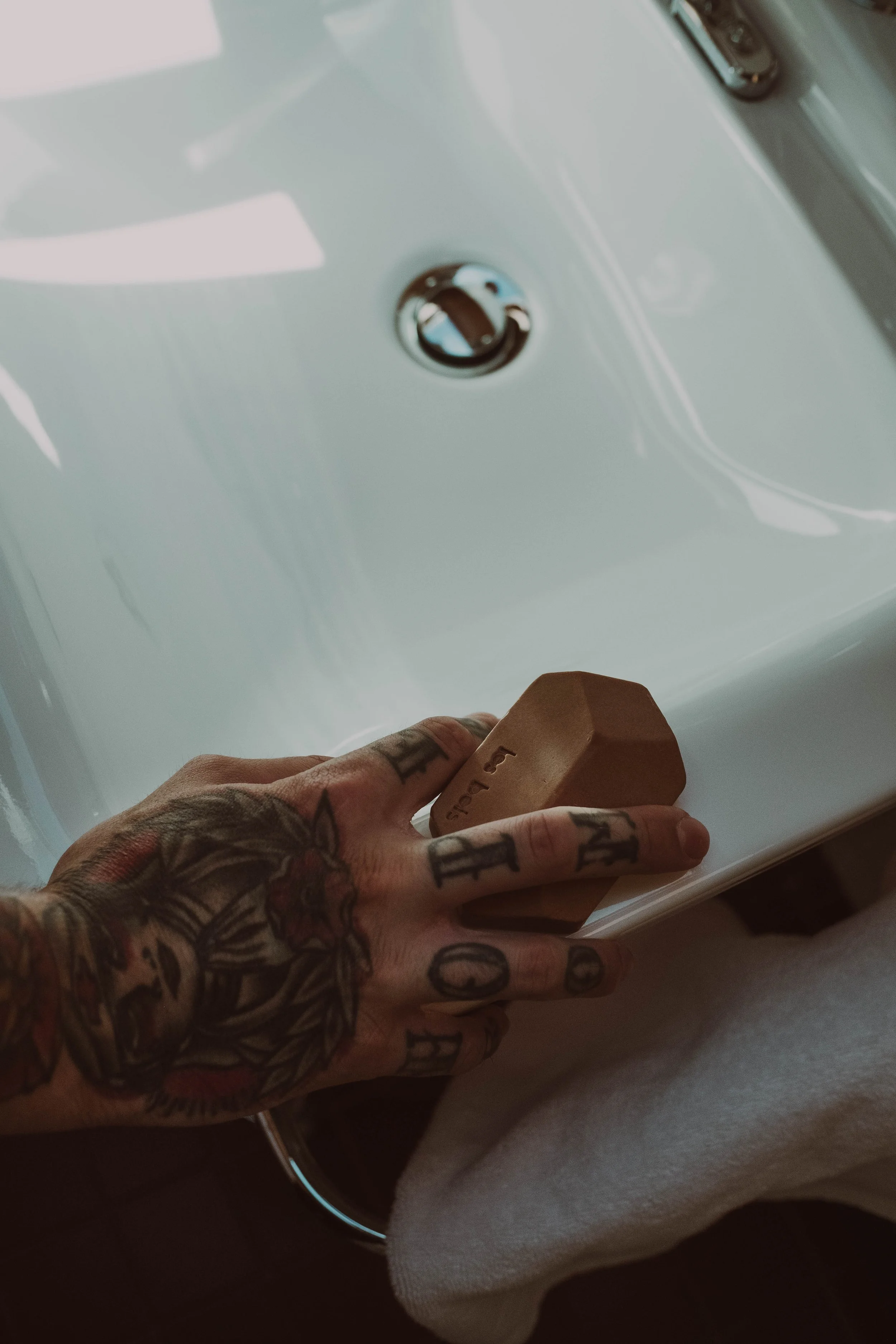 Une main tatouée tenant une éponge en bois, près d'un lavabo blanc.