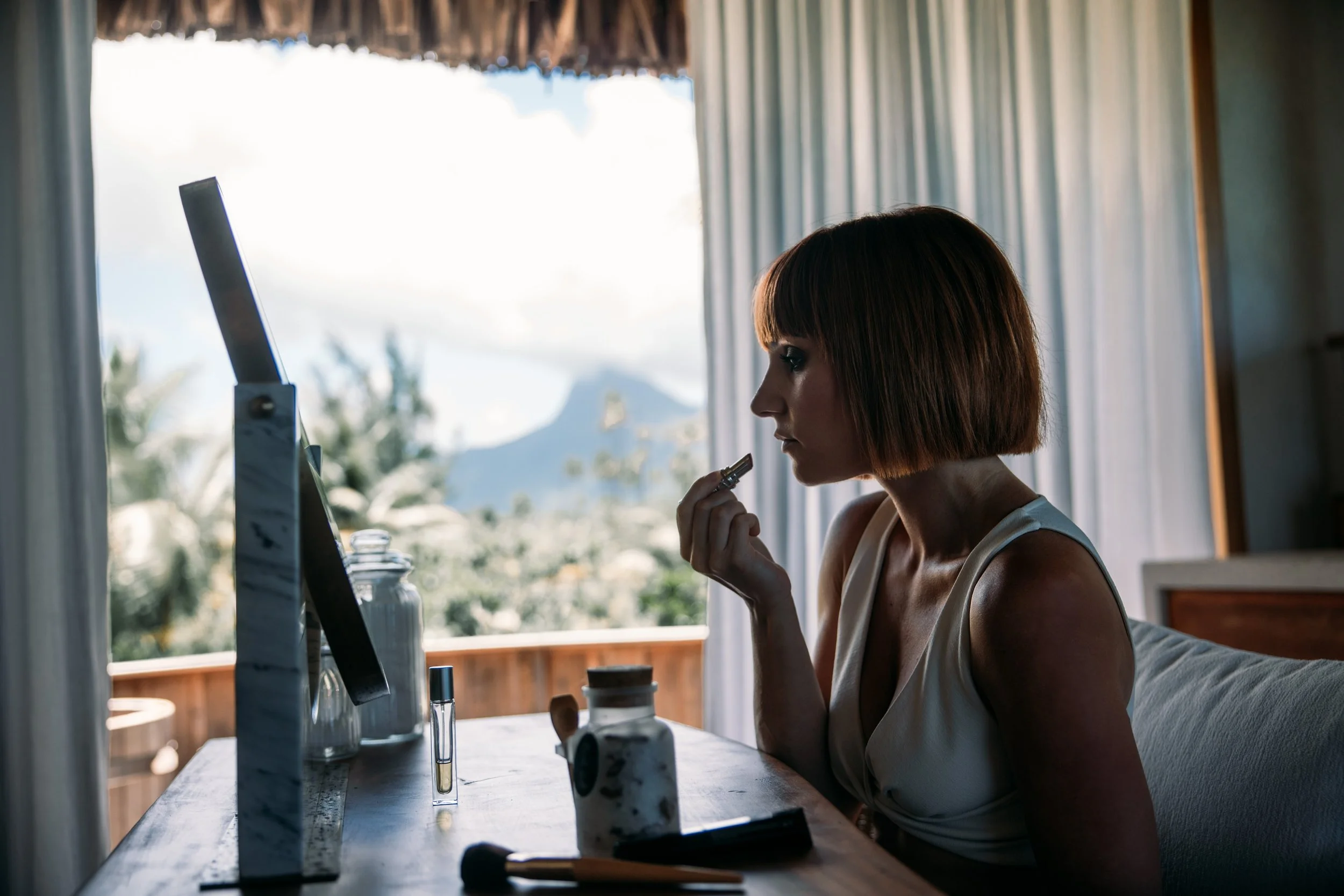 Une femme assise à une table près d'une fenêtre, regardant un miroir et appliquant du maquillage, avec un paysage naturel en arrière-plan.