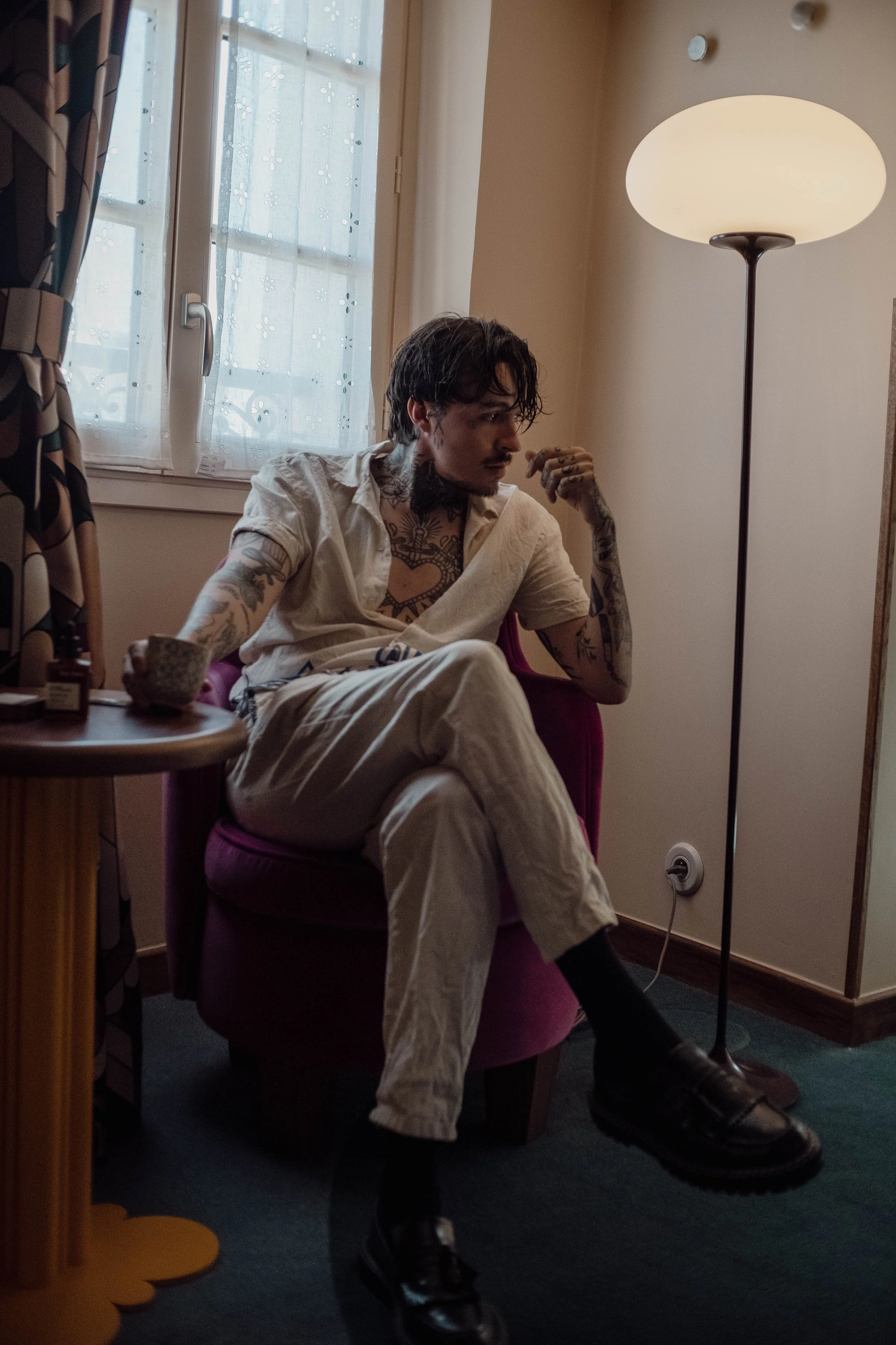 Un homme assis dans un fauteuil rose, portant une chemise beige et un pantalon beige, avec des tatouages visibles sur les bras et la poitrine, dans une pièce avec un lampadaire et un fenêtre. Il regarde pensivement.