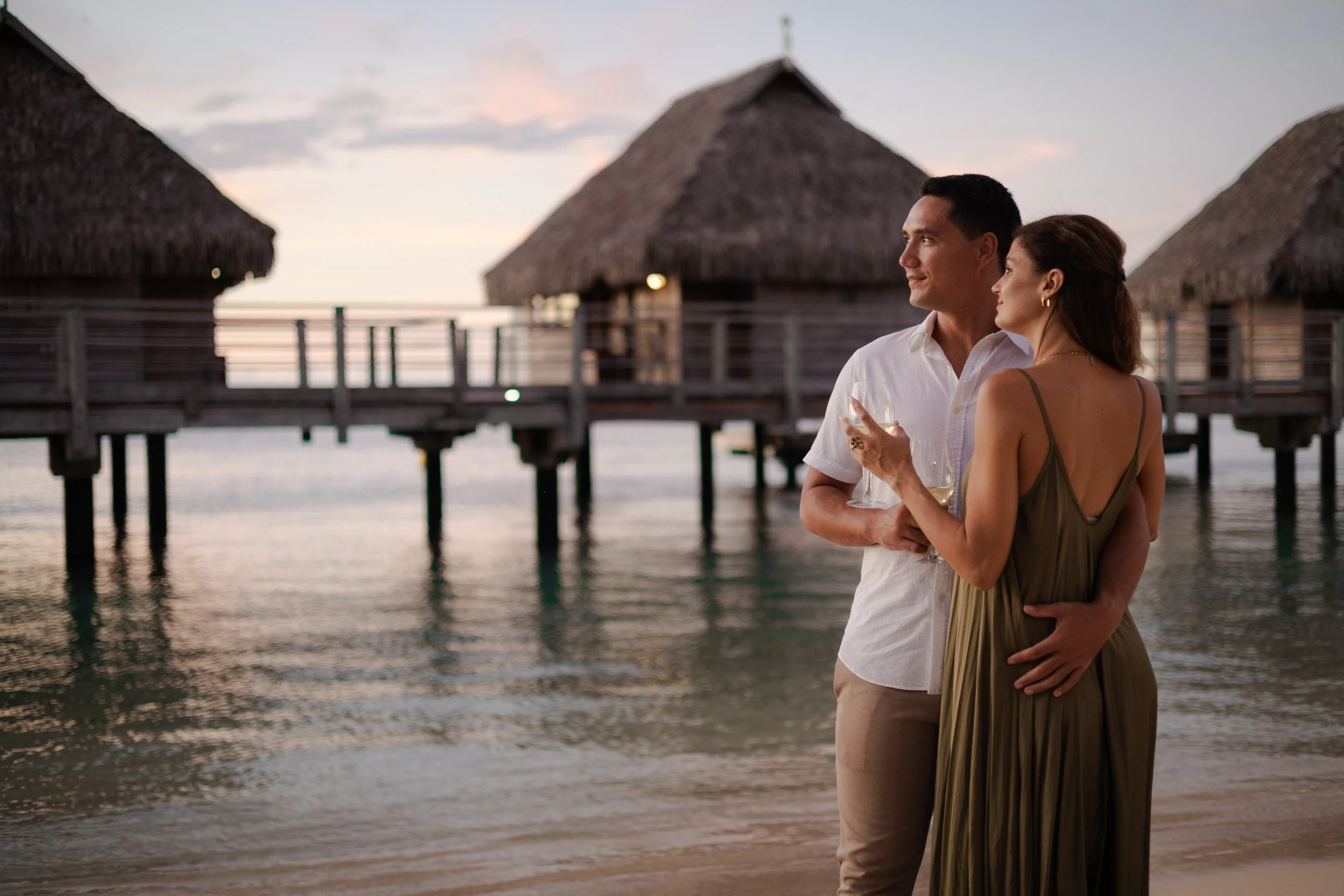 Un couple qui profite d'une soirée romantique sur une plage avec des huttes en bois sur pilotis en arrière-plan, au coucher du soleil.