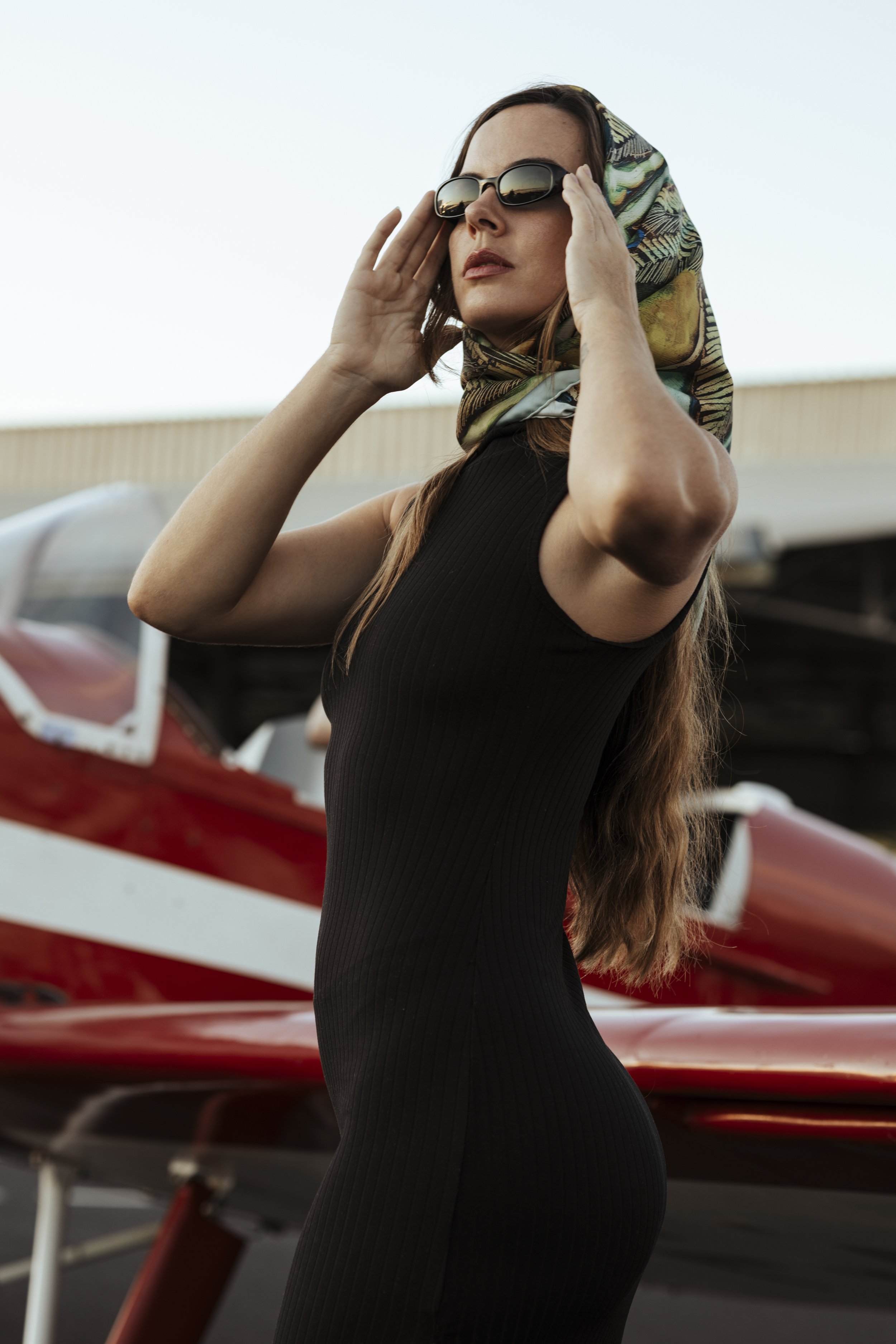Une femme portant des lunettes de soleil, un foulard coloré sur la tête, en tenue noire, se tenant à côté d'un avion rouge.