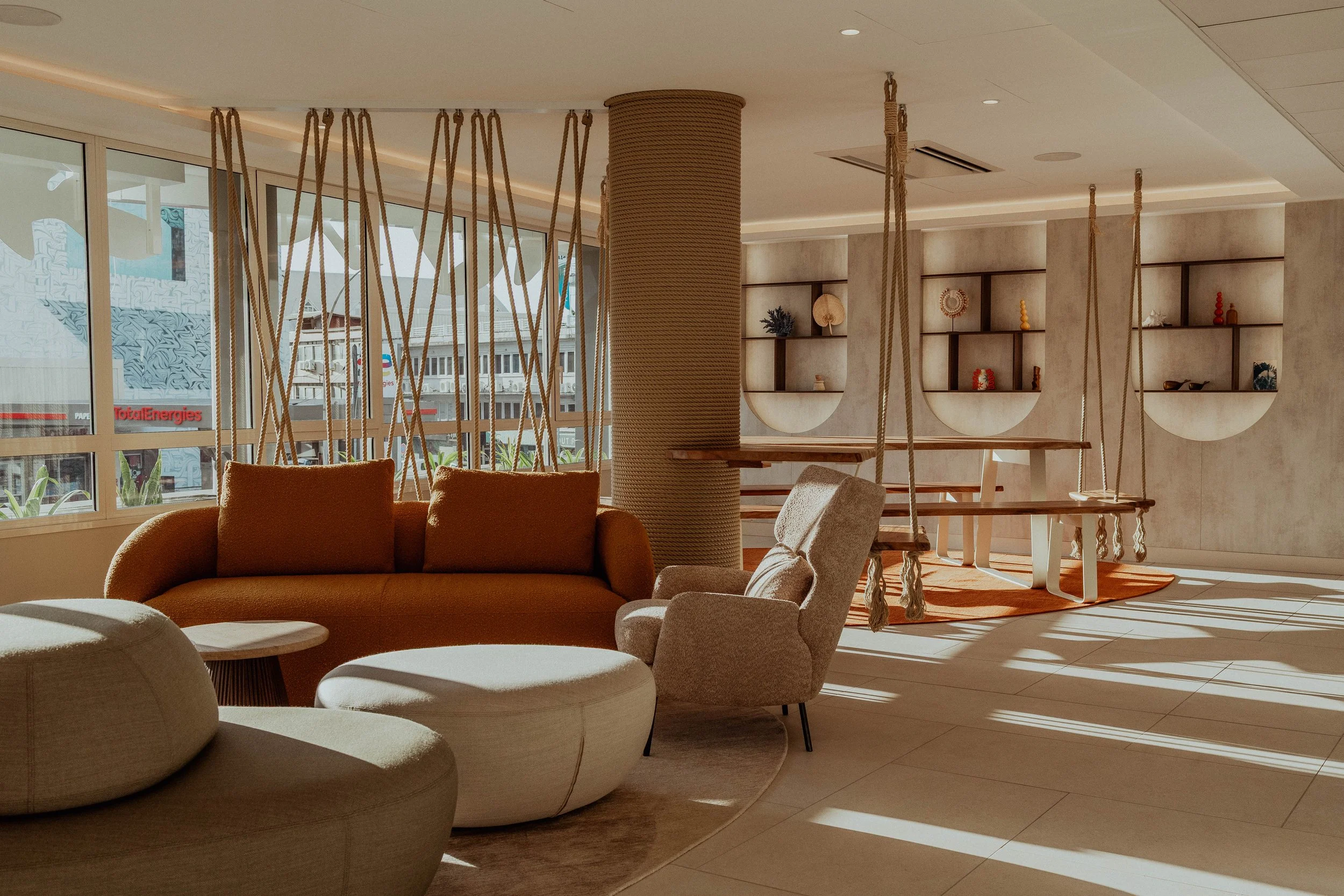 Salon moderne avec canapé beige, fauteuil gris, pouf blanc et balançoires en corde suspendues devant de grandes fenêtres lumineuses.