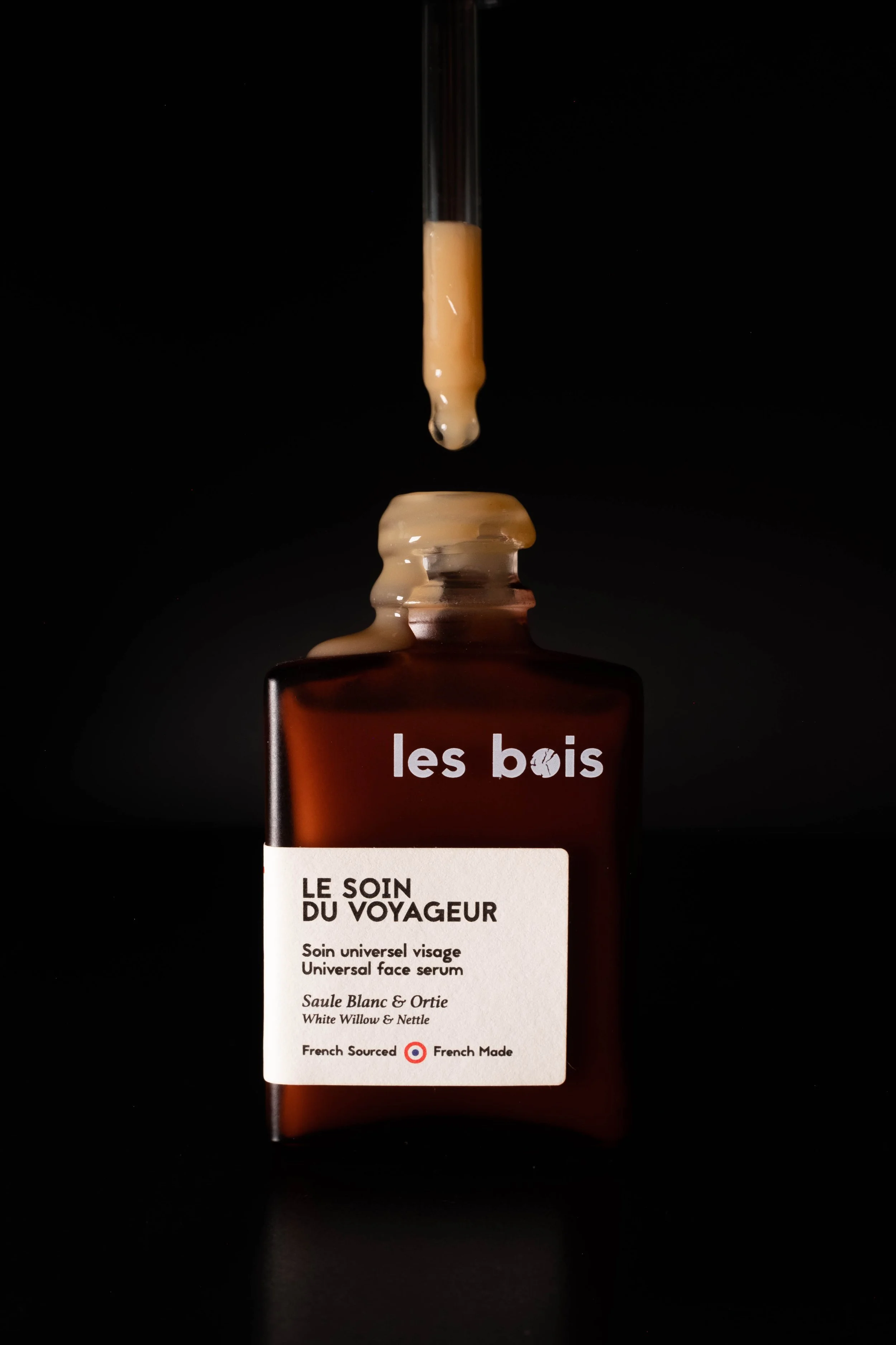 Une bouteille de sérum visage brun avec un pipette déversant du sérum. L'étiquette indique 'les bois' et 'LE SOIN DU VOYAGEUR', avec des détails en français et en anglais.