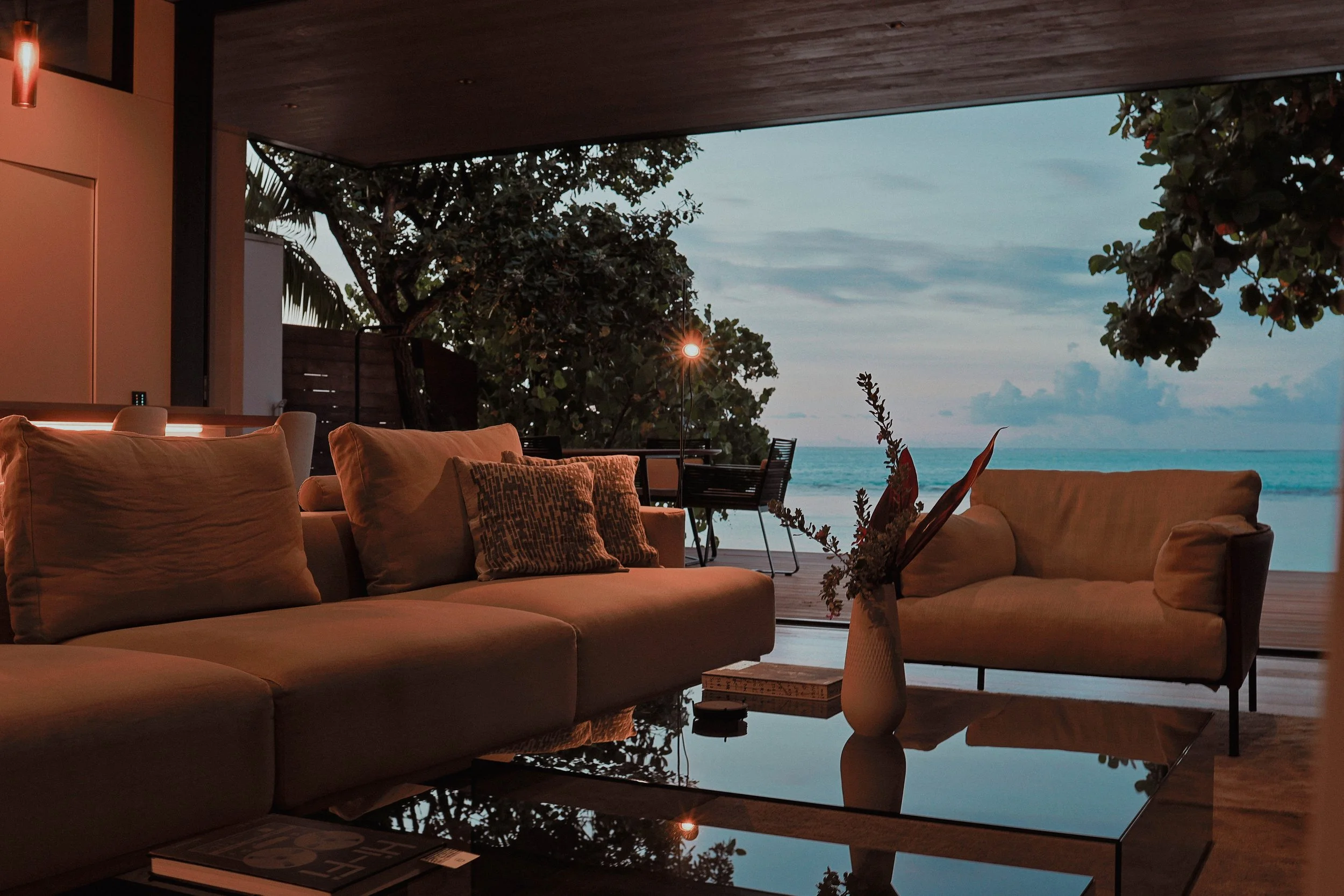 Salon intérieur moderne avec canapé et fauteuils, vue sur la mer à travers une grande ouverture, vase avec des fleurs, table basse en verre.