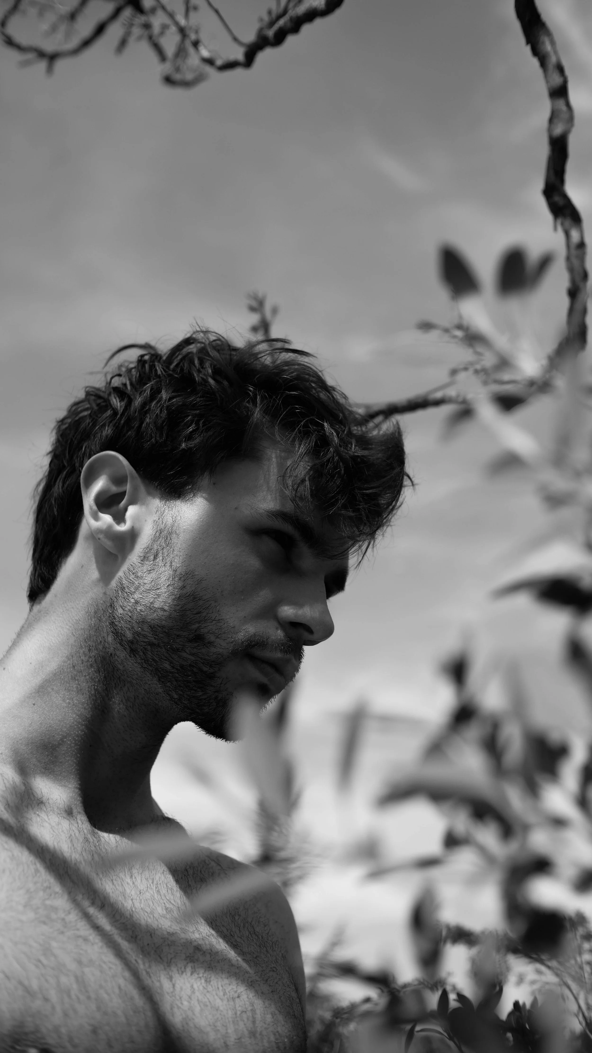 Jeune homme non habillé, avec barbe, regardant vers le bas, dans un champ avec des plantes en noir et blanc.
