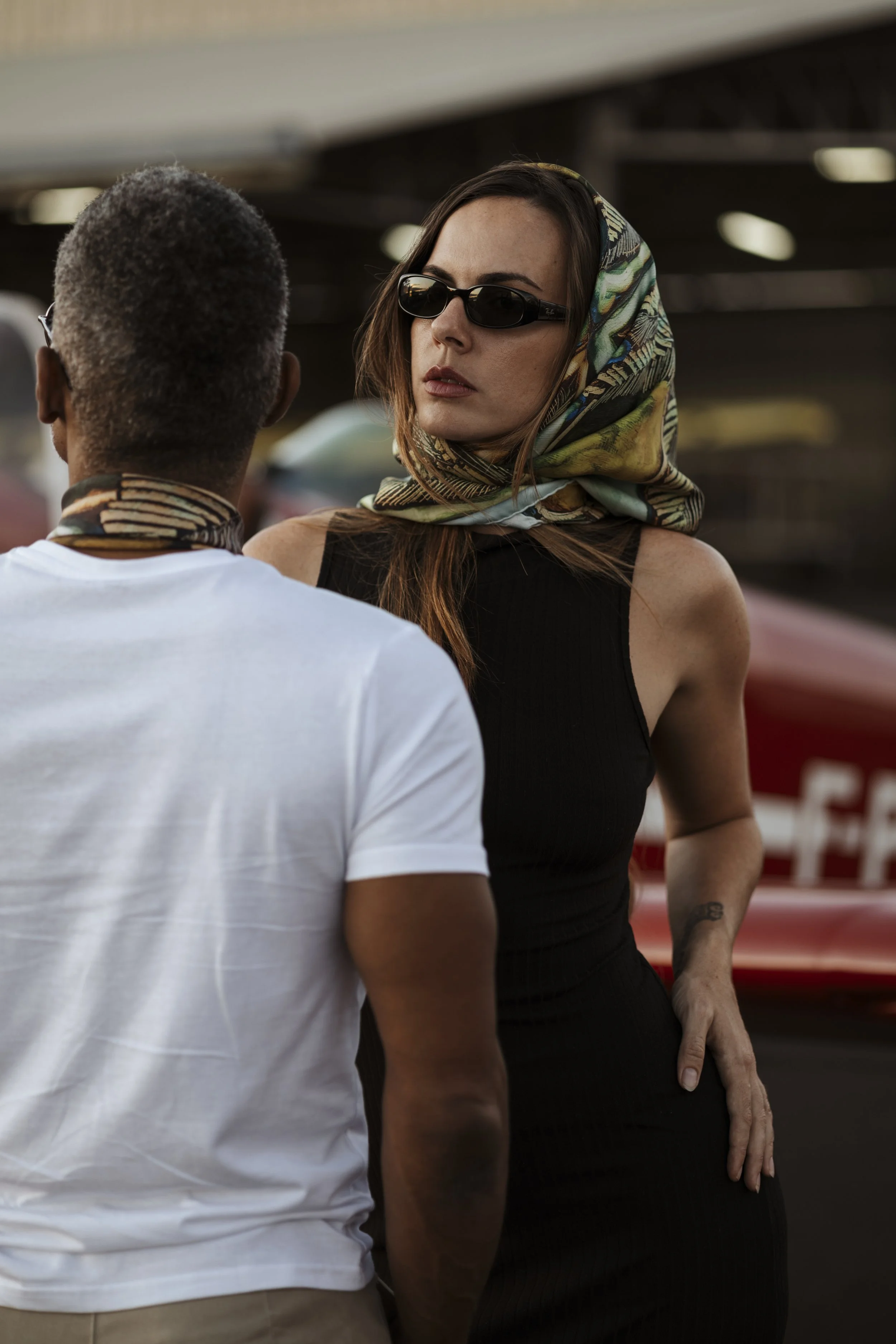 Une femme portant des lunettes de soleil noires, un foulard coloré sur la tête et un gilet sans manches noir, discute avec un homme dont le dos et la tête sont visibles, portant un t-shirt blanc et un foulard autour du cou, près d'une voiture rouge.