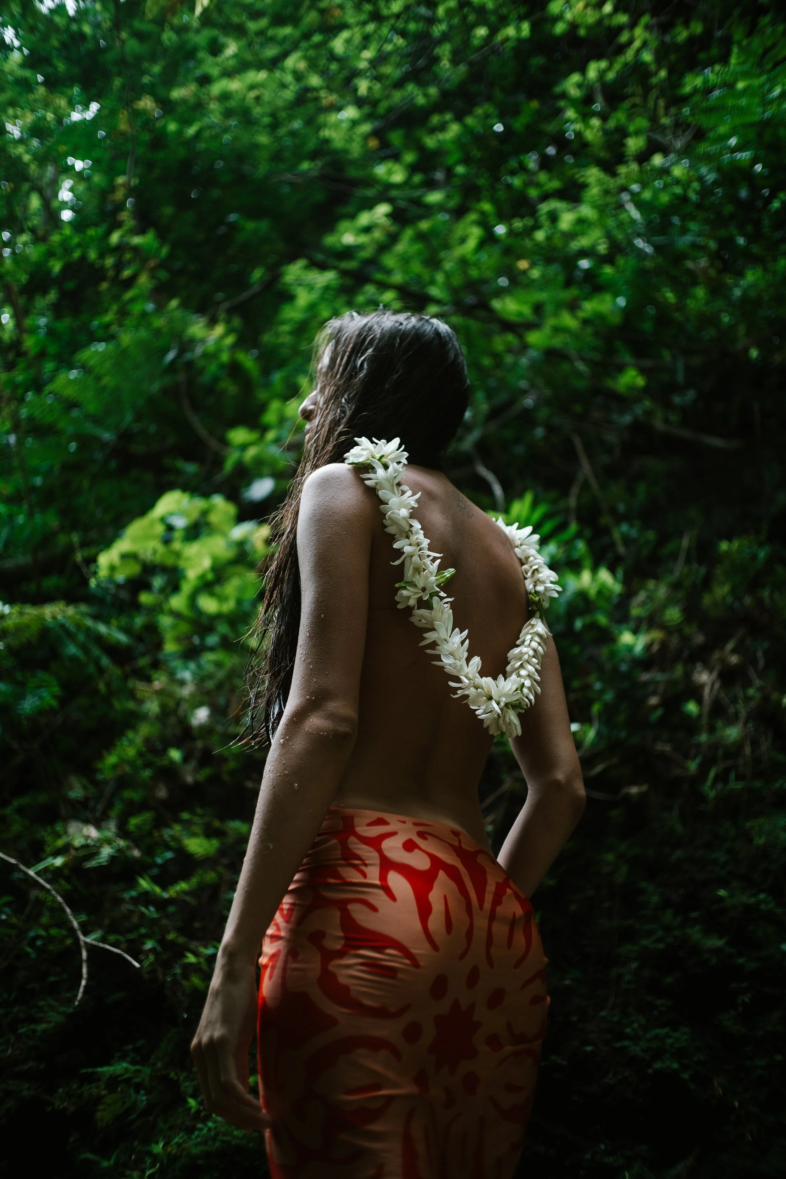 Femme nue portant une guirlande de fleurs et une jupe orange avec des motifs rouges, dans une forêt dense et verte.