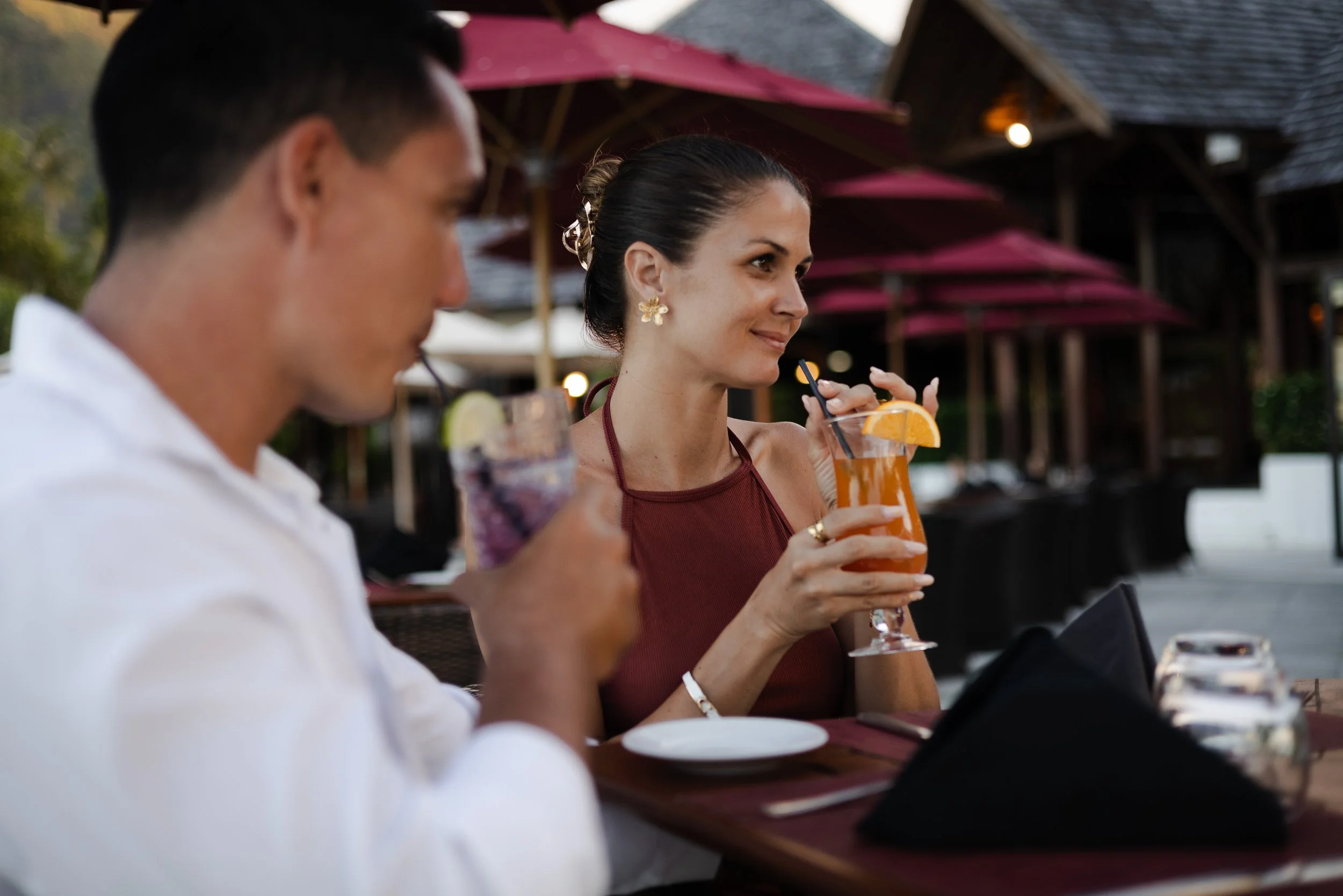 Deux personnes assises à une table de restaurant en plein air, dégustant des boissons colorées avec des décorations d'agrumes, dans un cadre extérieur avec parasols et bâtiments en bois.