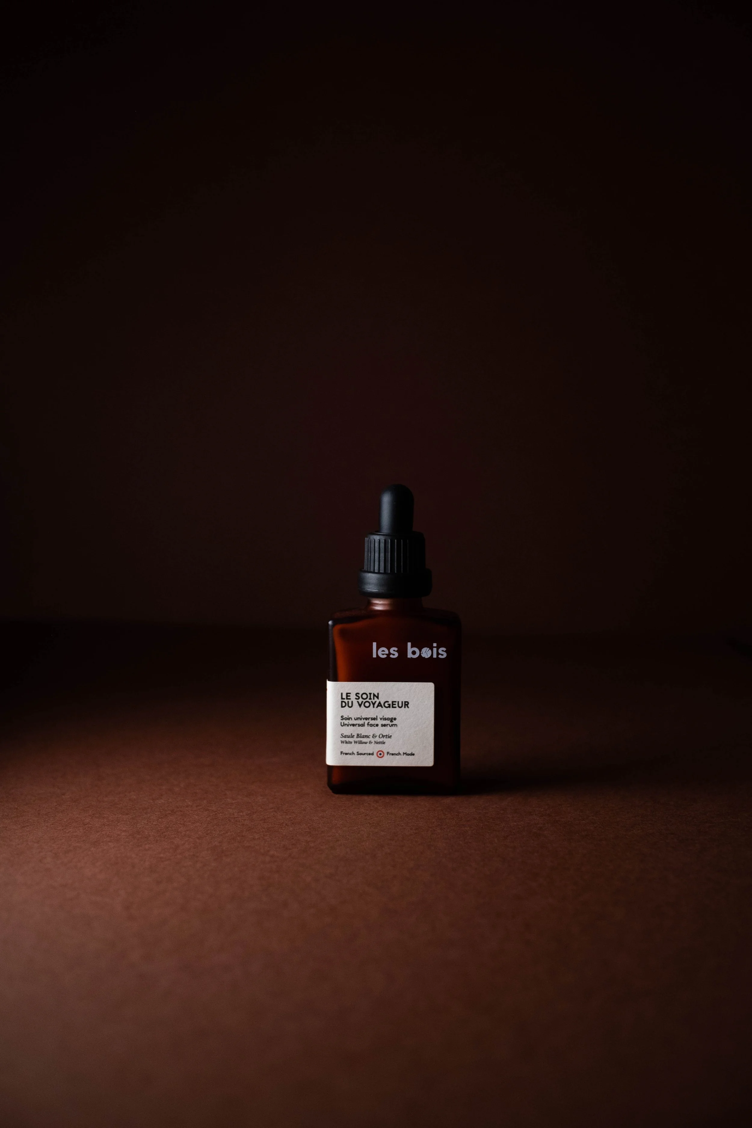Une bouteille en verre marron avec un compte-gouttes, étiquetée 'les bois' et 'LE SOIN DU VOYAGEUR', posée sur une surface marron foncé avec un fond sombre et une lumière douce. 