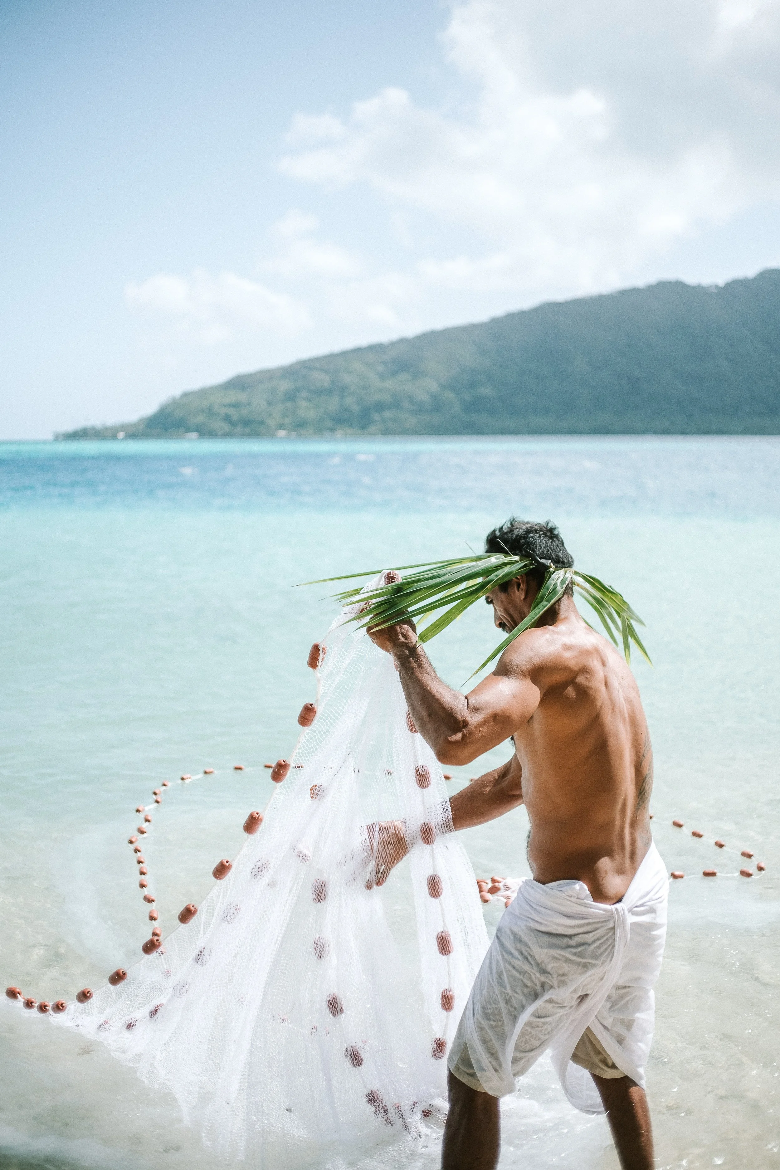 Hommes pêchant à la plage avec un filet de pêche, portant des feuilles de palmier sur la tête, en arrière-plan de la mer et une île ou une montagne verte.