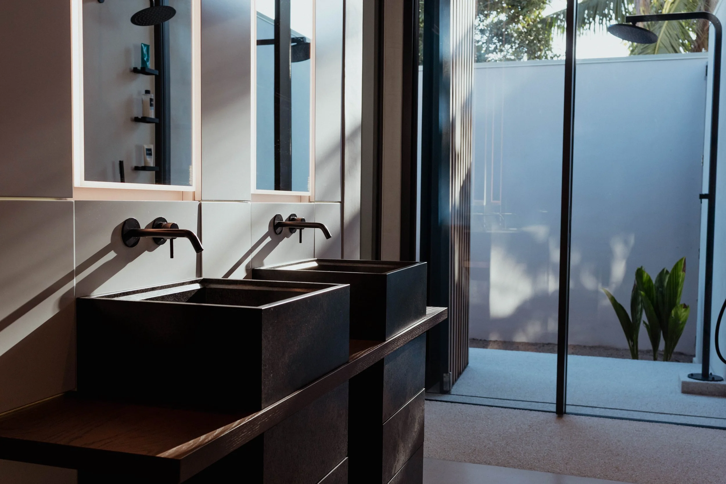 Lavabos noirs avec robinets noirs dans une salle de bain moderniste, avec miroirs et un espace extérieur visible à travers une porte en verre.