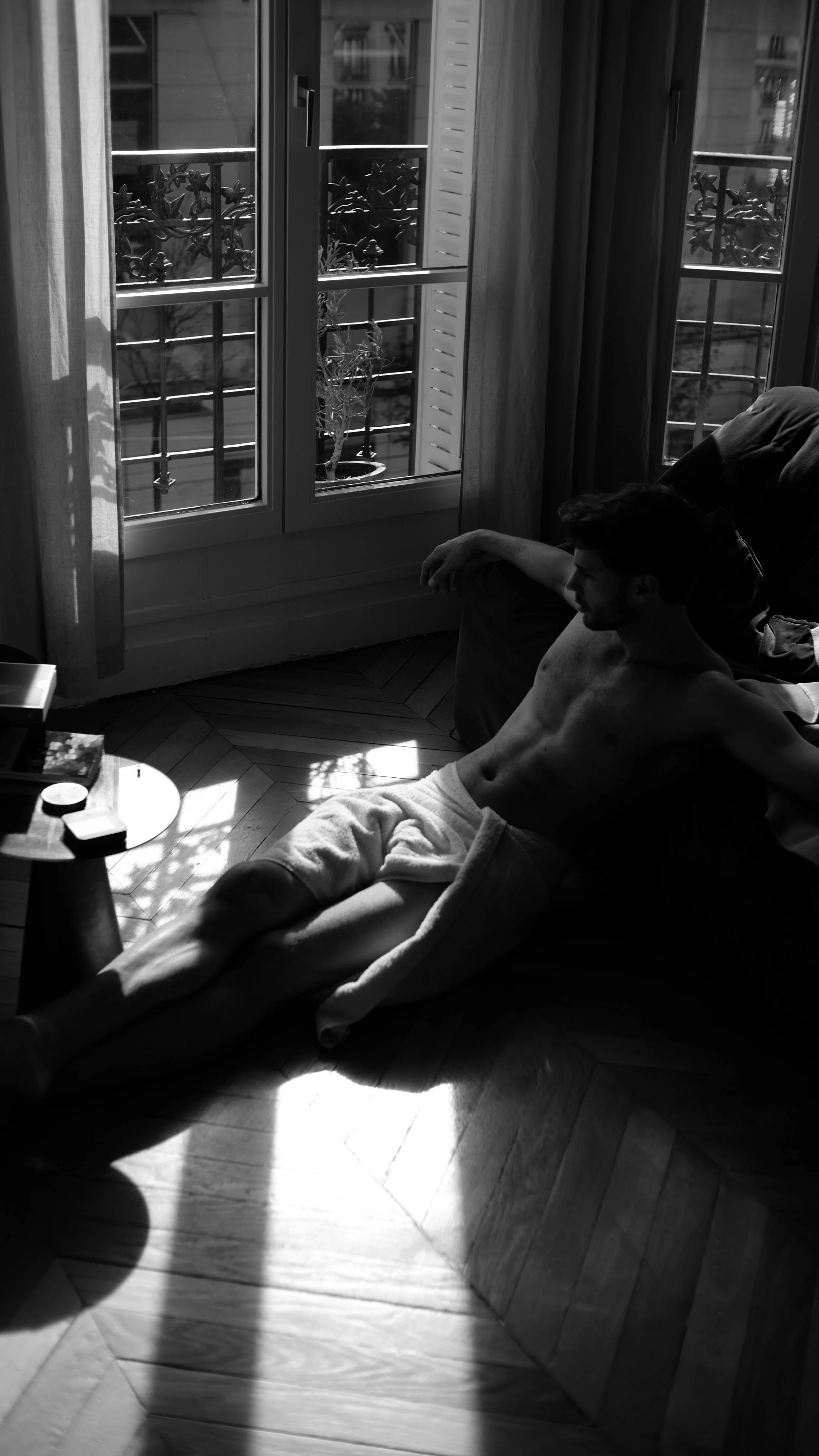 Un homme allongé sur un canapé, dans une pièce avec de grandes fenêtres laissant entrer la lumière du soleil, en noir et blanc.