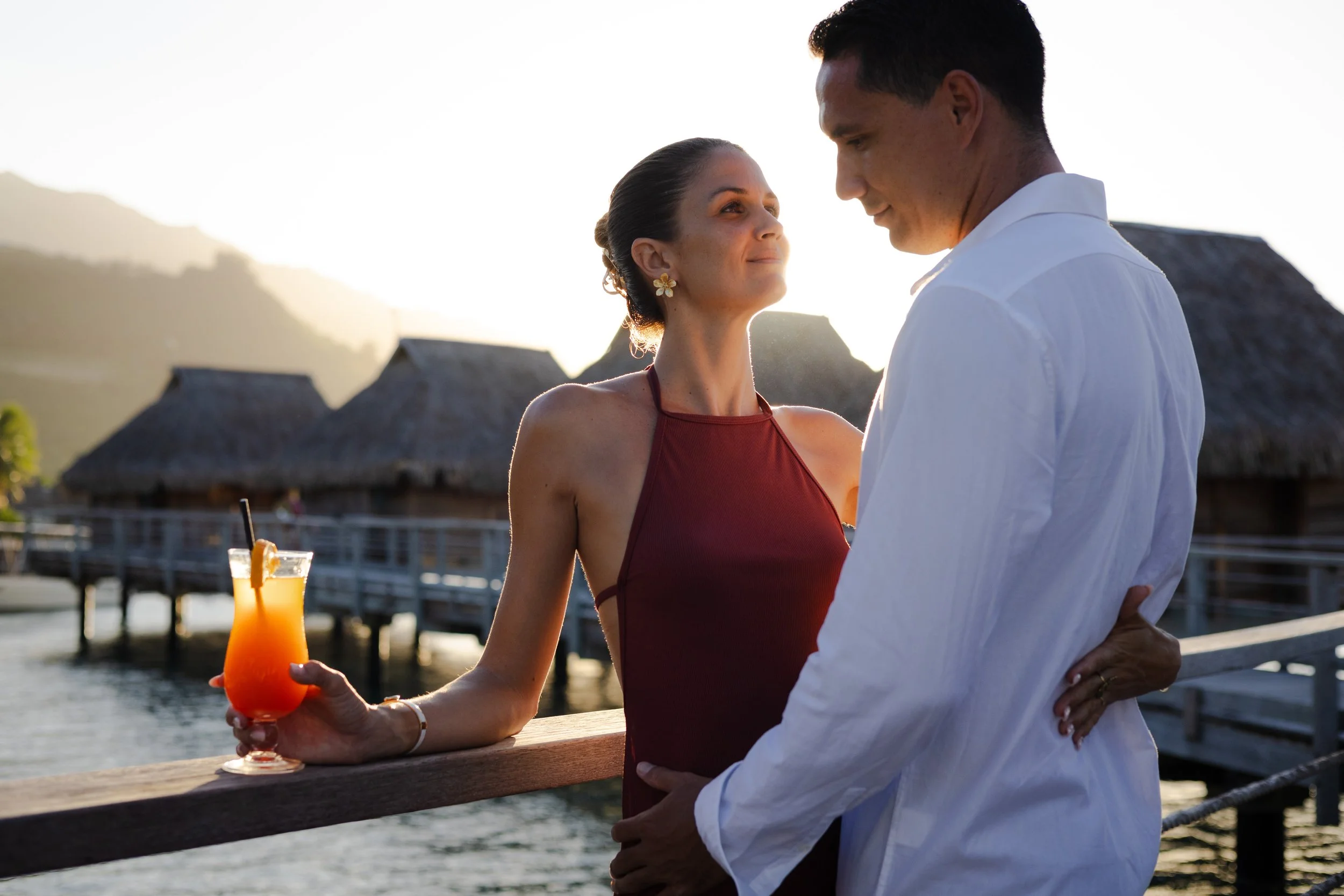 Un couple tente une embrassade près d'une jetée en soirée, avec un fond de cabanes en bois sur l'eau et un paysage de montagnes, la femme tient un cocktail coloré.