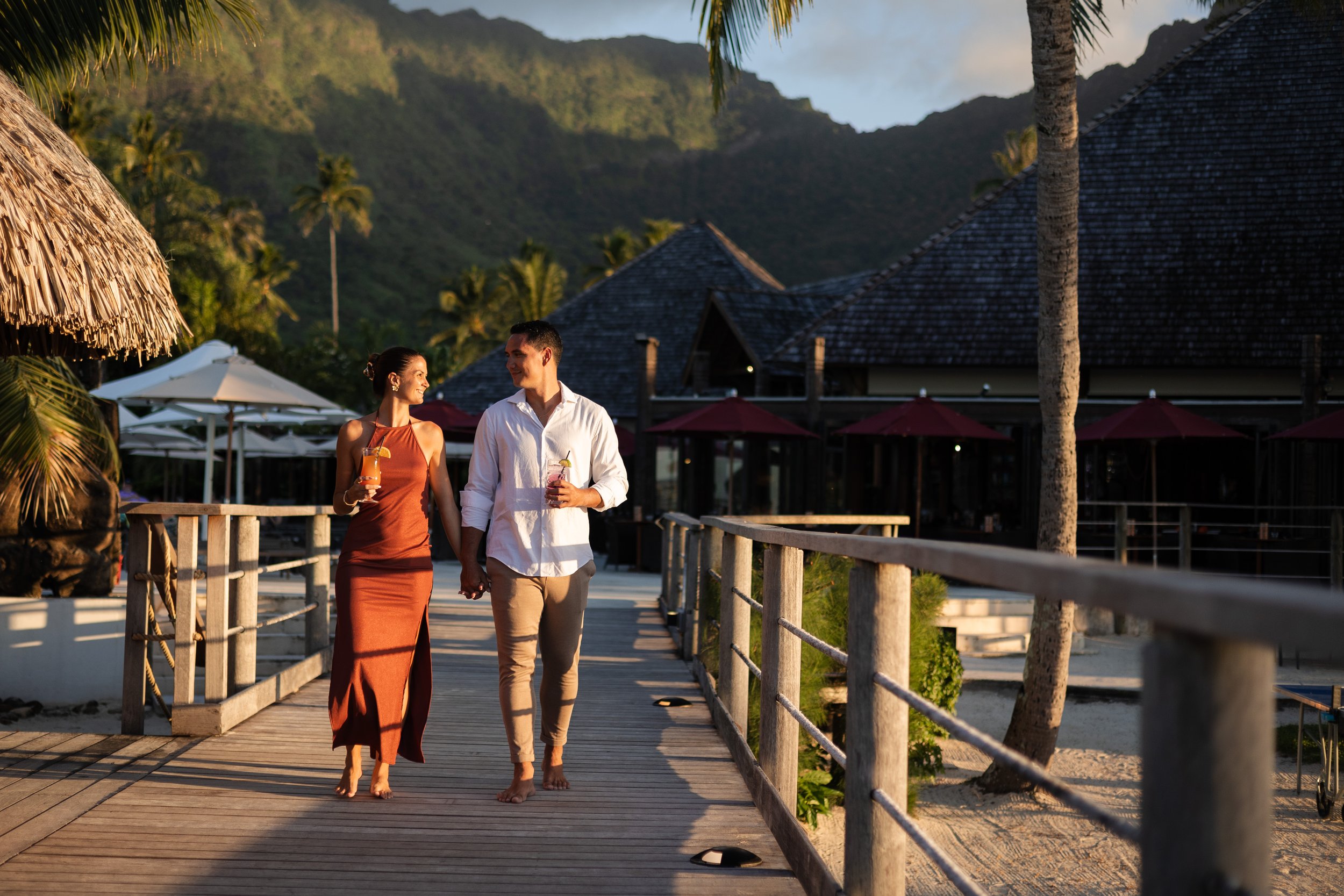Un couple marchant main dans la main sur une jetée en bois, tenant des boissons dans chaque main, avec un décor tropical comprenant des palmiers et des bâtiments avec des toits en bois dans un environnement ensoleillé et montagneux.