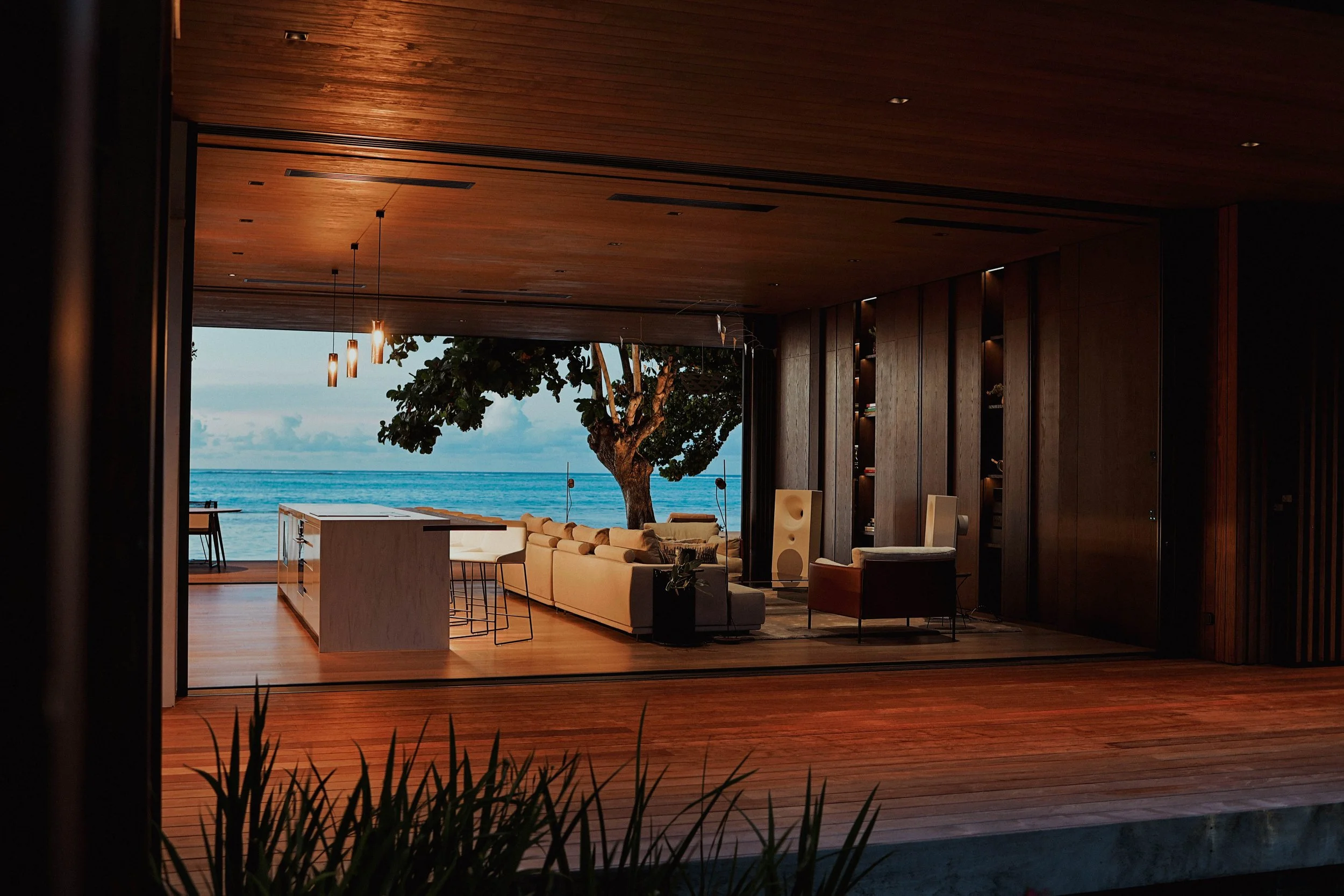 Intérieur d'une maison moderne avec vue sur la mer, canapé beige, salon avec mobilier en bois, éclairage tamisé, arbre visible à travers une grande ouverture.
