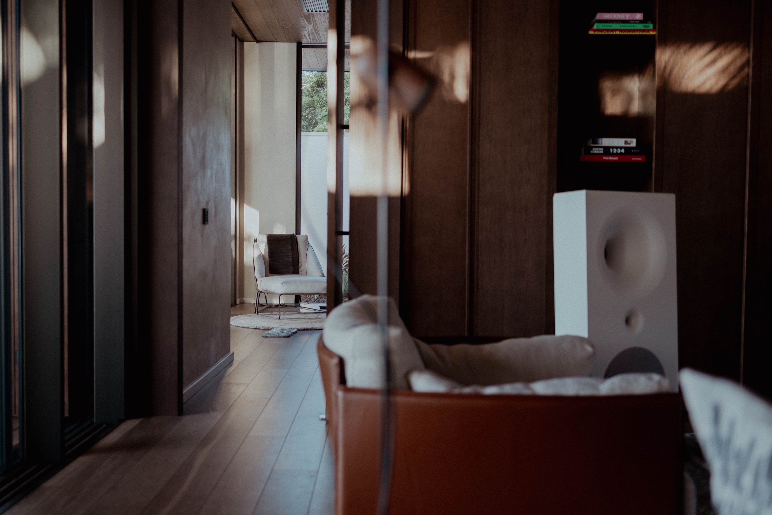 Vue intérieure d'un salon avec un fauteuil blanc en arrière-plan, un canapé beige en avant-plan, une enceinte voilée blanche, et une étagère avec des livres, dans une pièce avec des murs en bois et un sol en bois.