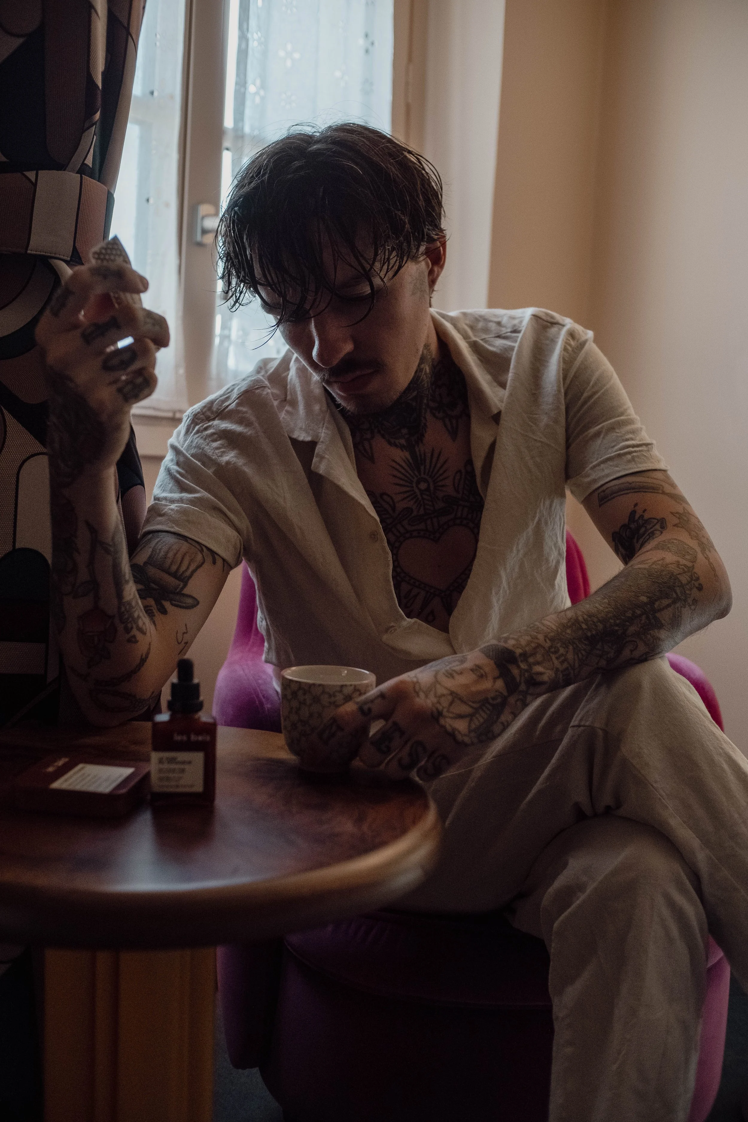 Un homme tatoué, assis à une table, tenant une tasse, avec des médicaments sur la table et un fond de fenêtre avec rideau.
