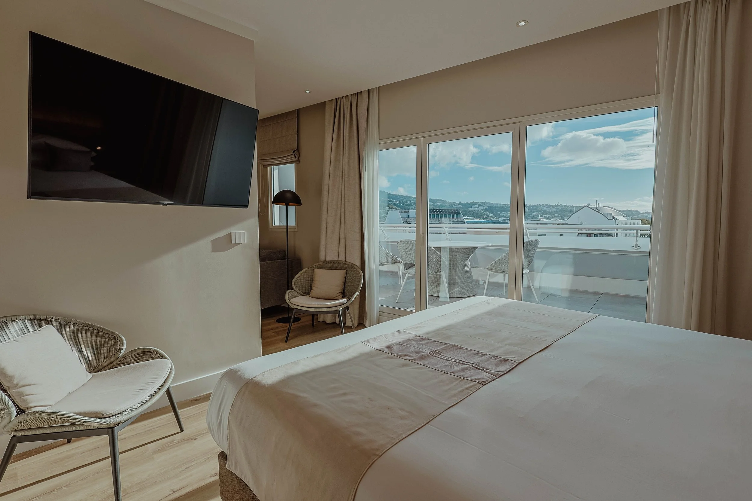 Chambre d'hôtel avec un lit, un fauteuil, un petit canapé, un téléviseur mural et une terrasse avec vue sur la ville et le ciel.