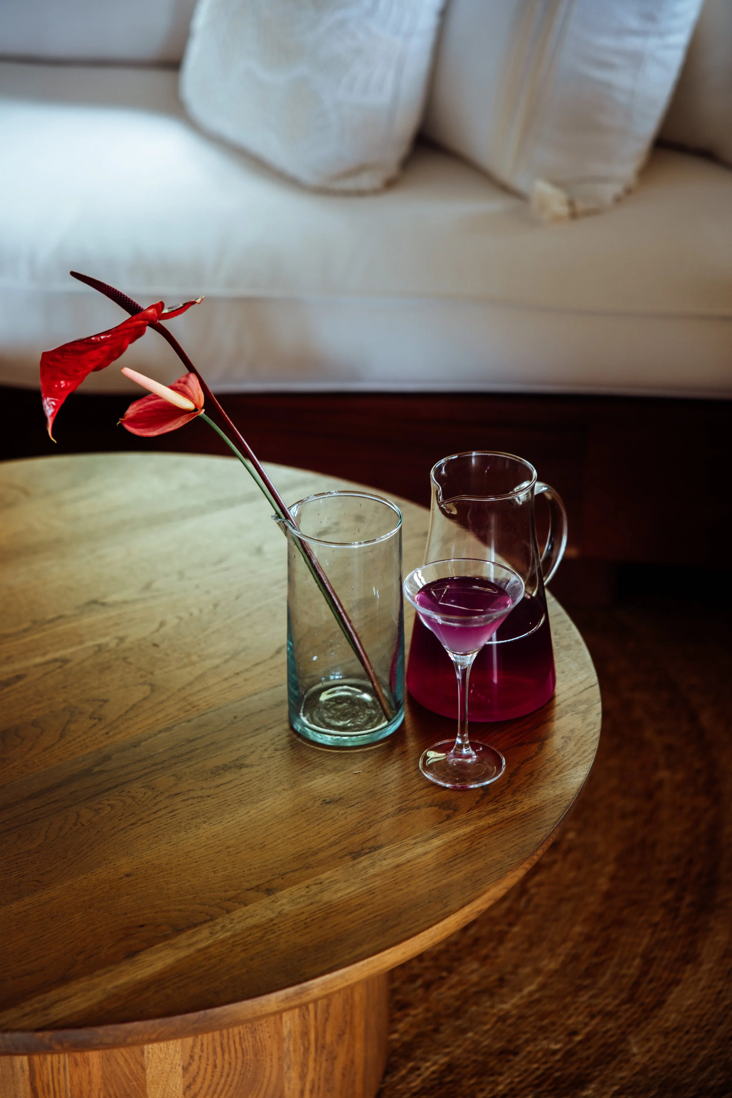 Un verre transparent avec une touche de fleurs rouges, un pichet rempli d'un liquide violet foncé, et un petit verre à cocktail violet clair, tous placés sur une table en bois près d'un canapé beige avec des coussins
