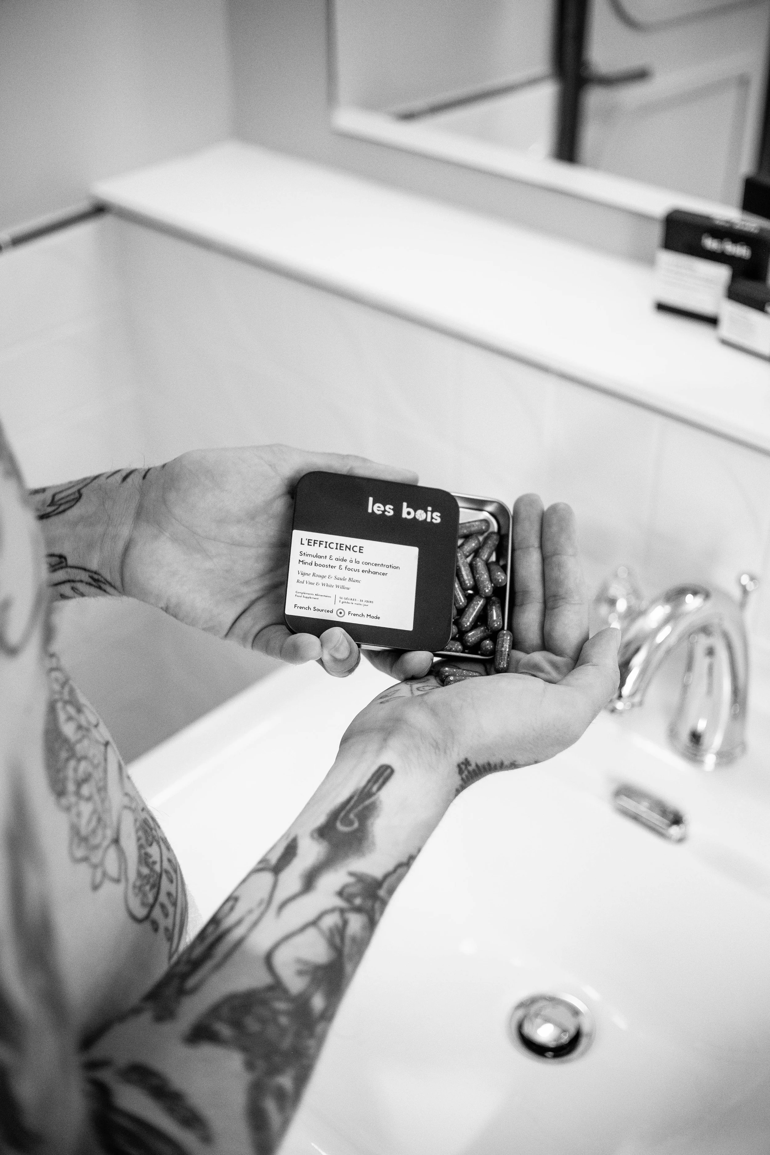 Une personne tenant un petit contenant en métal avec des capsules, marqué "les bois" à l'extérieur, dans une salle de bain, avec une baignoire blanche, un miroir et des boîtes à l'arrière-plan.