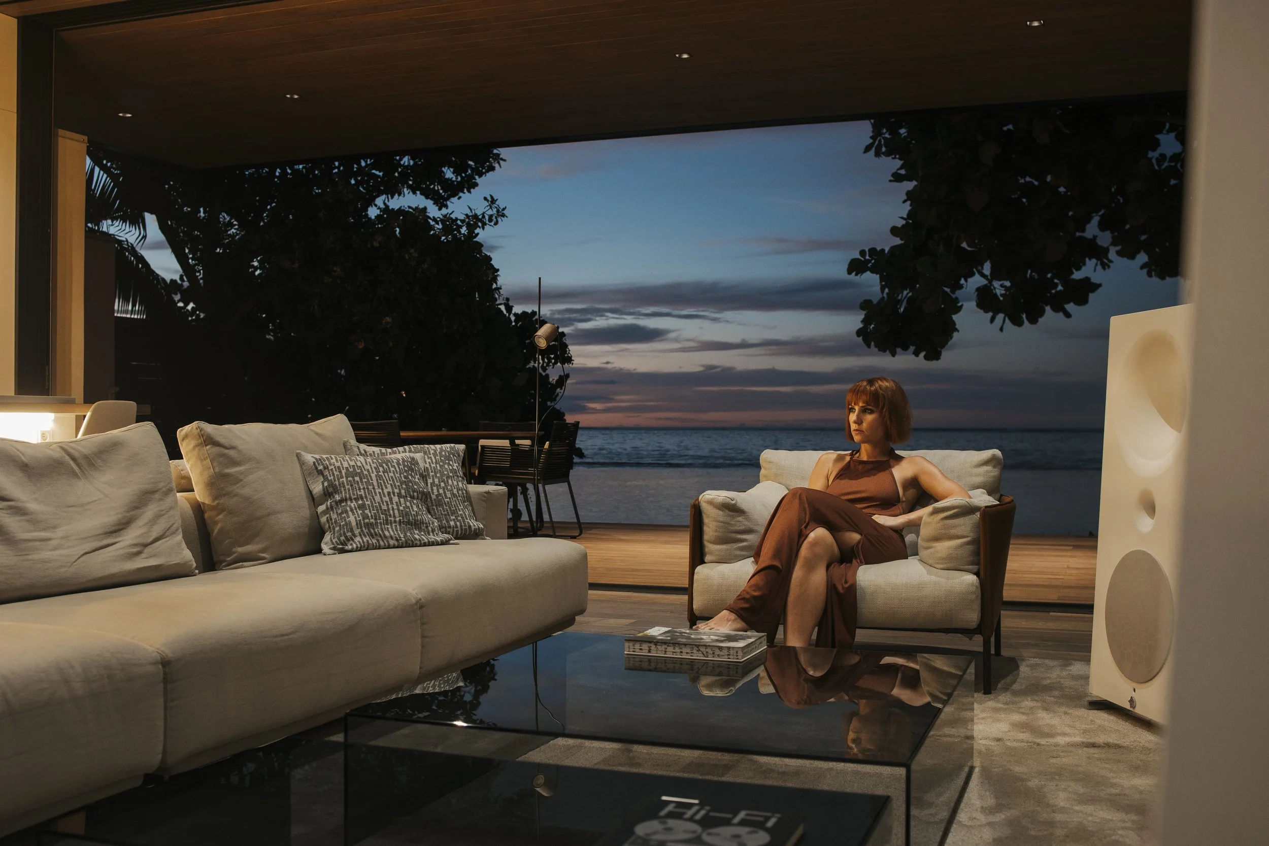 Une femme assise sur un canapé dans un salon près de l'océan au coucher du soleil, avec vue sur la mer et les arbres en arrière-plan.