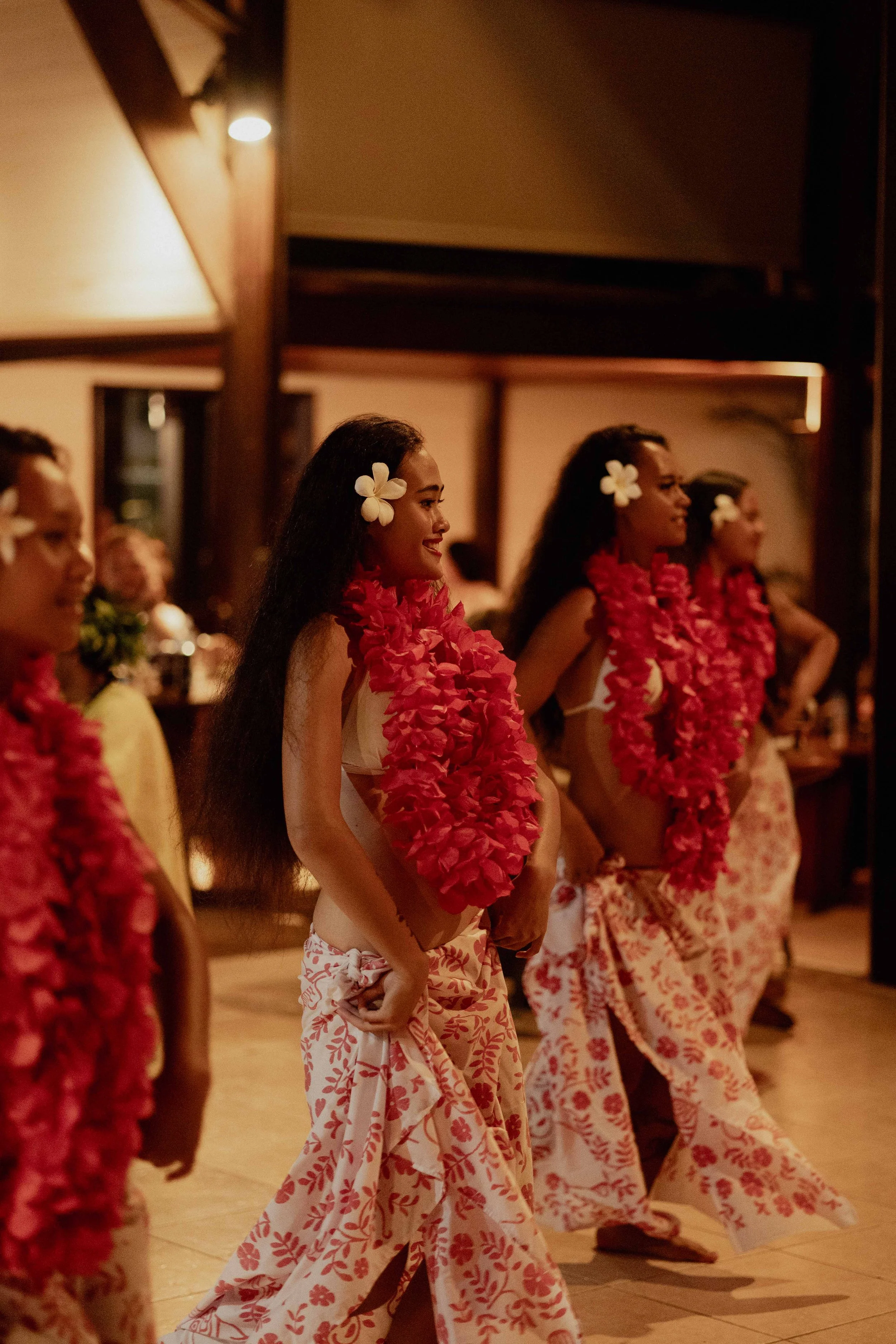 Femmes en costume traditionnel hawaïen, portant des colliers de fleurs roses et des jupes à motifs rouges et blancs, participant à une danse polynésienne dans une salle chaleureuse.