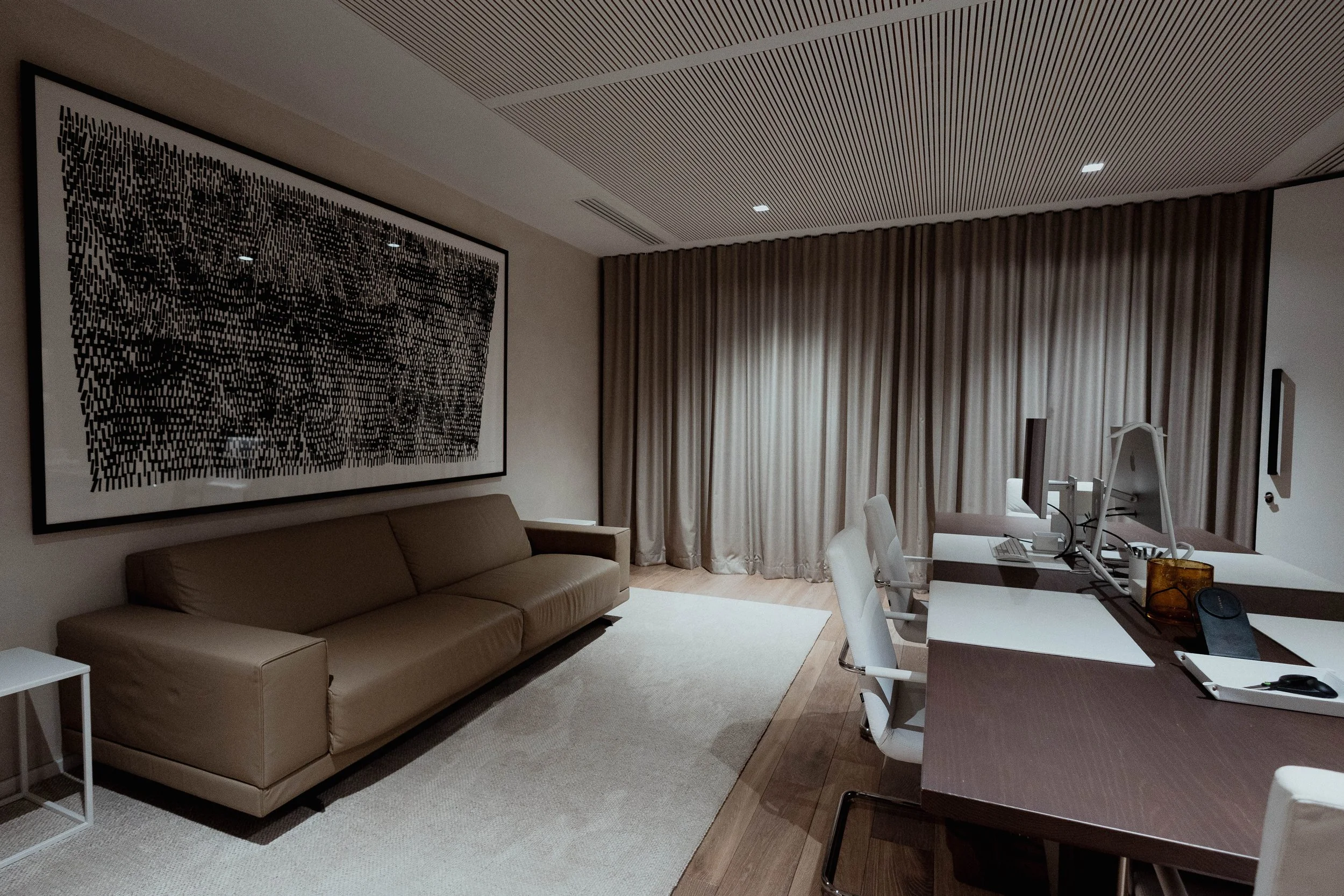 Salon moderne avec un canapé beige, une grande œuvre d'art noire et blanche sur le mur, une grande table de conférence avec des ordinateurs et des chaises blanches, et de longs rideaux gris clairs en arrière-plan.