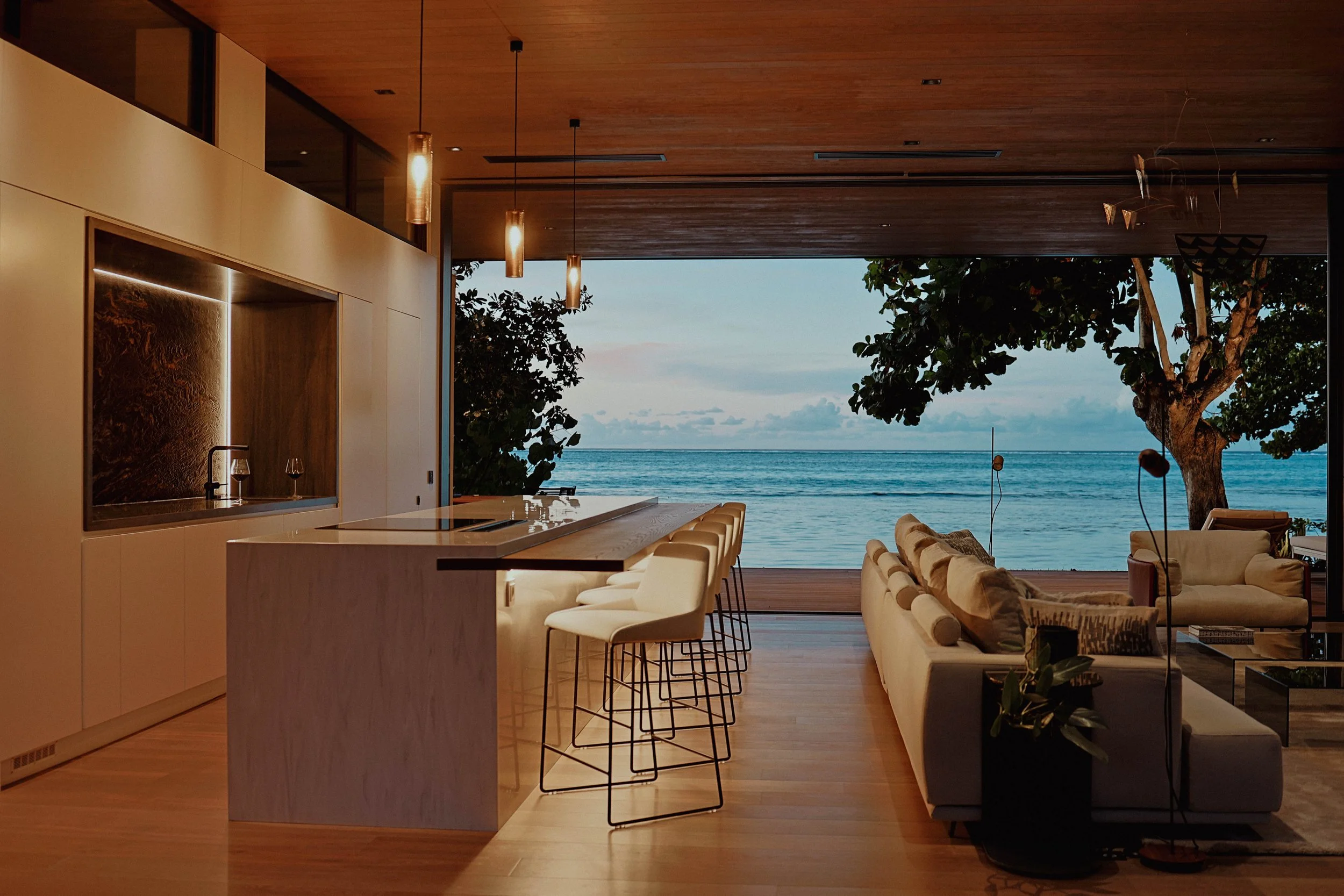 Intérieur d'une maison moderne avec vue sur la mer, comprenant un bar, une cuisine, un coin salon avec canapé, et un grand écran, avec une vue sur la mer et un arbre à l'extérieur.