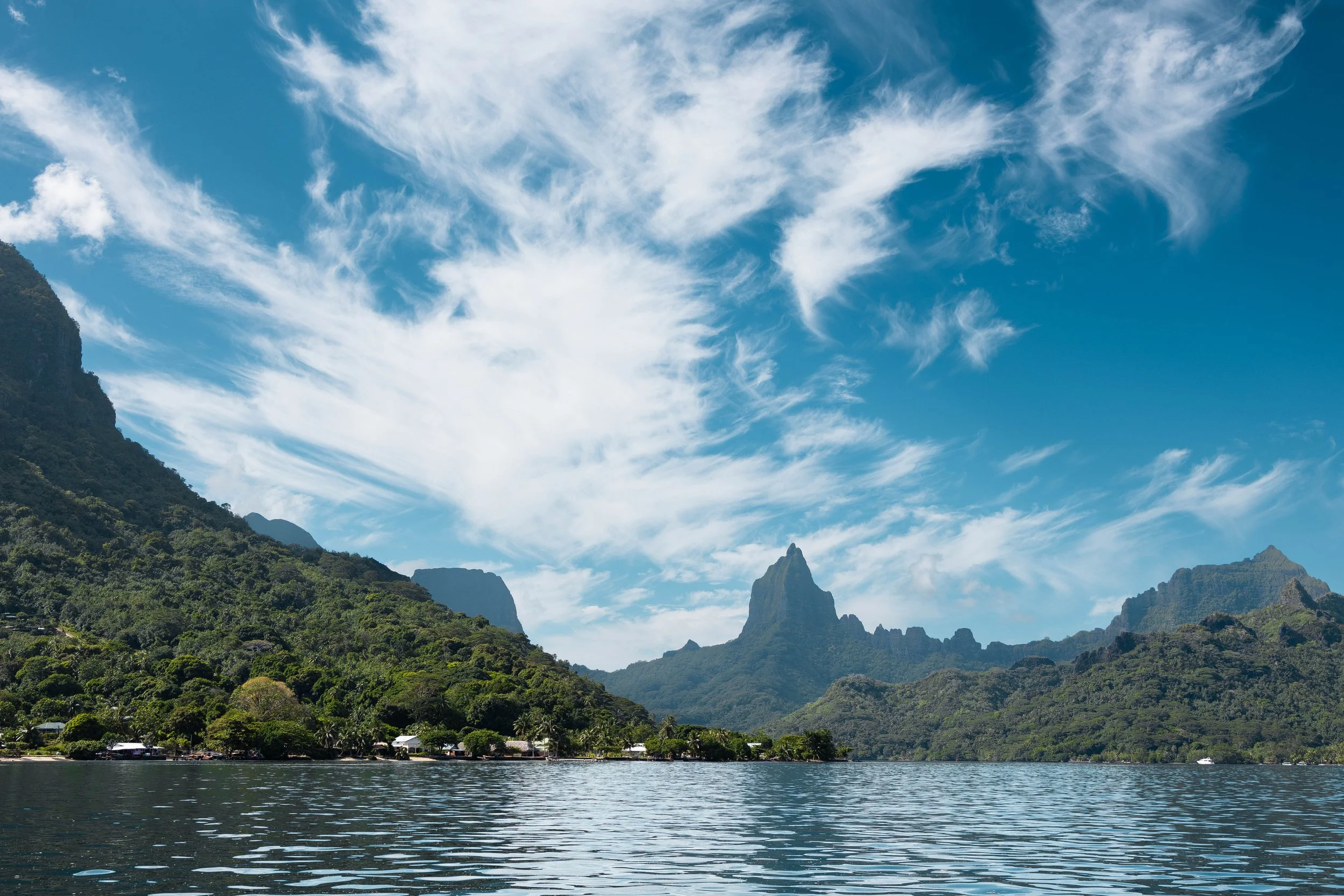 Paysage de montagnes verdoyantes avec une rivière ou un lac au premier plan, ciel bleu avec quelques nuages blancs.