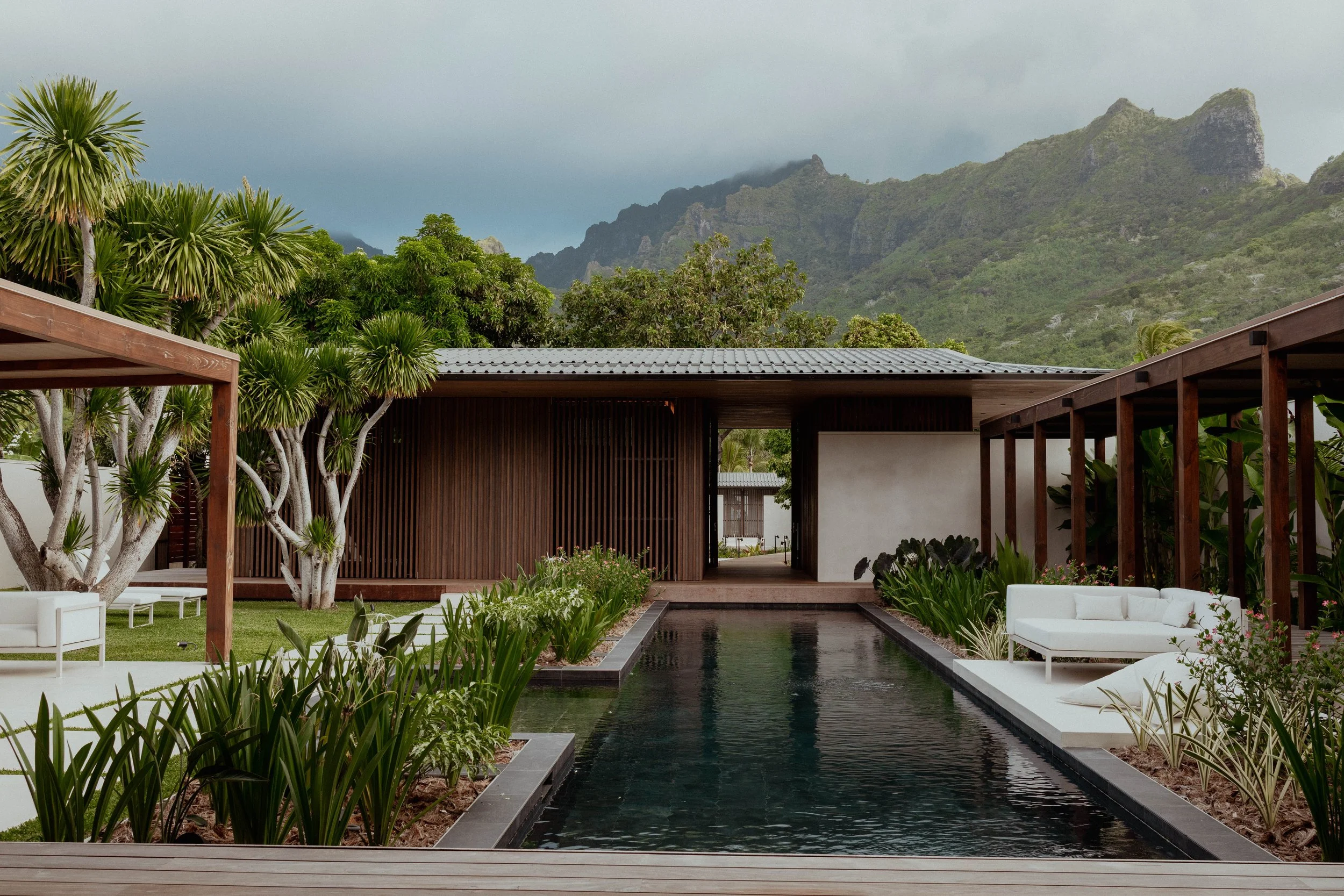 Un espace de détente avec une piscine noire au centre, entourée de plantes vertes, avec des meubles blancs et une structure en bois, avec des montagnes en arrière-plan.