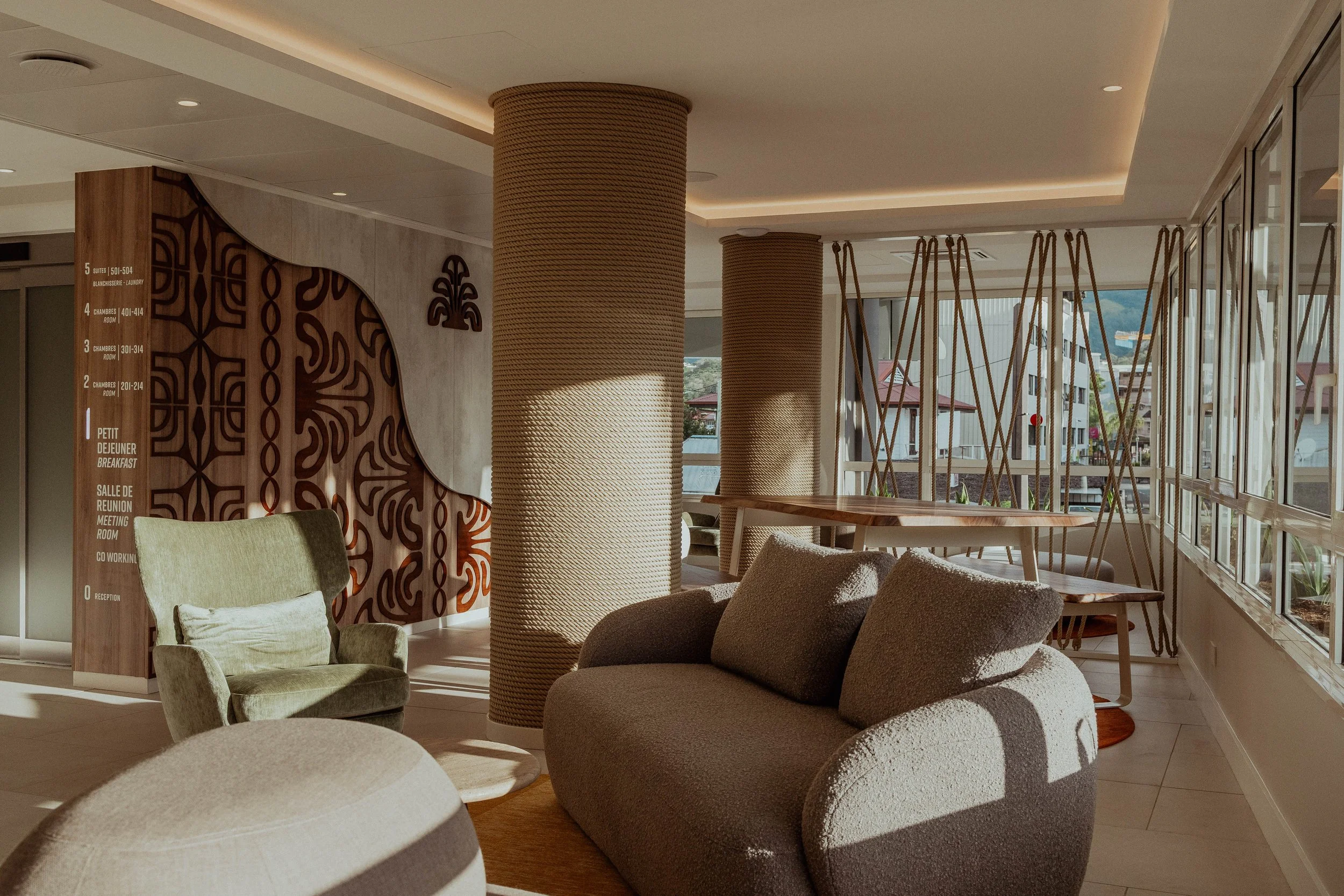 Salon lumineux avec canapé gris, fauteuil vert, colonnes recouvertes de corde, et une table en bois, avec des fenêtres offrant une vue sur des bâtiments.