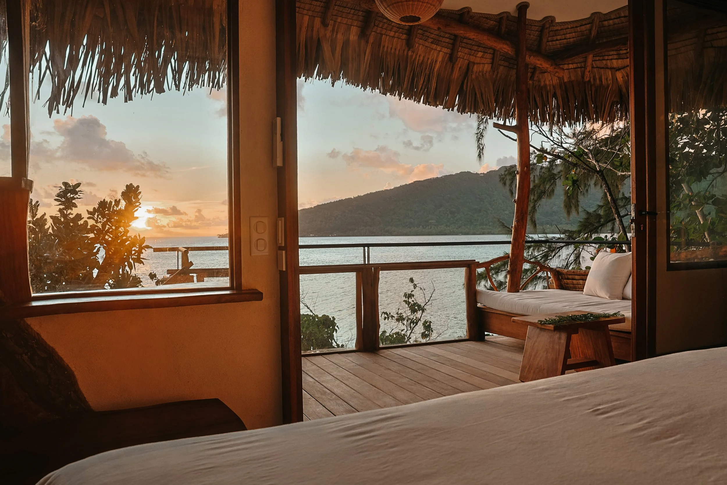 Vue d'une chambre avec balcon en bois, ouverte sur un paysage marin avec une île ou une montagne en arrière-plan, au coucher du soleil.