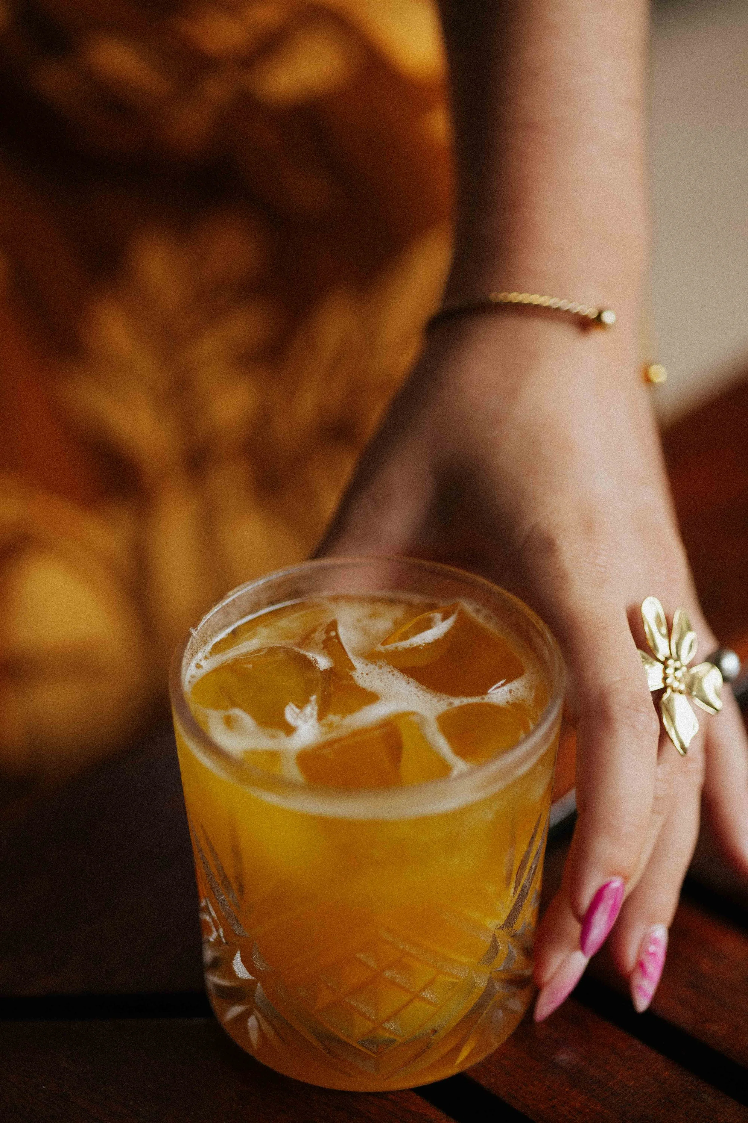 Une main féminine tenant un verre avec une boisson orange, des glaçons, et de la mousse, posée sur une surface en bois. La main porte des bijoux, dont un anneau en forme de fleur.