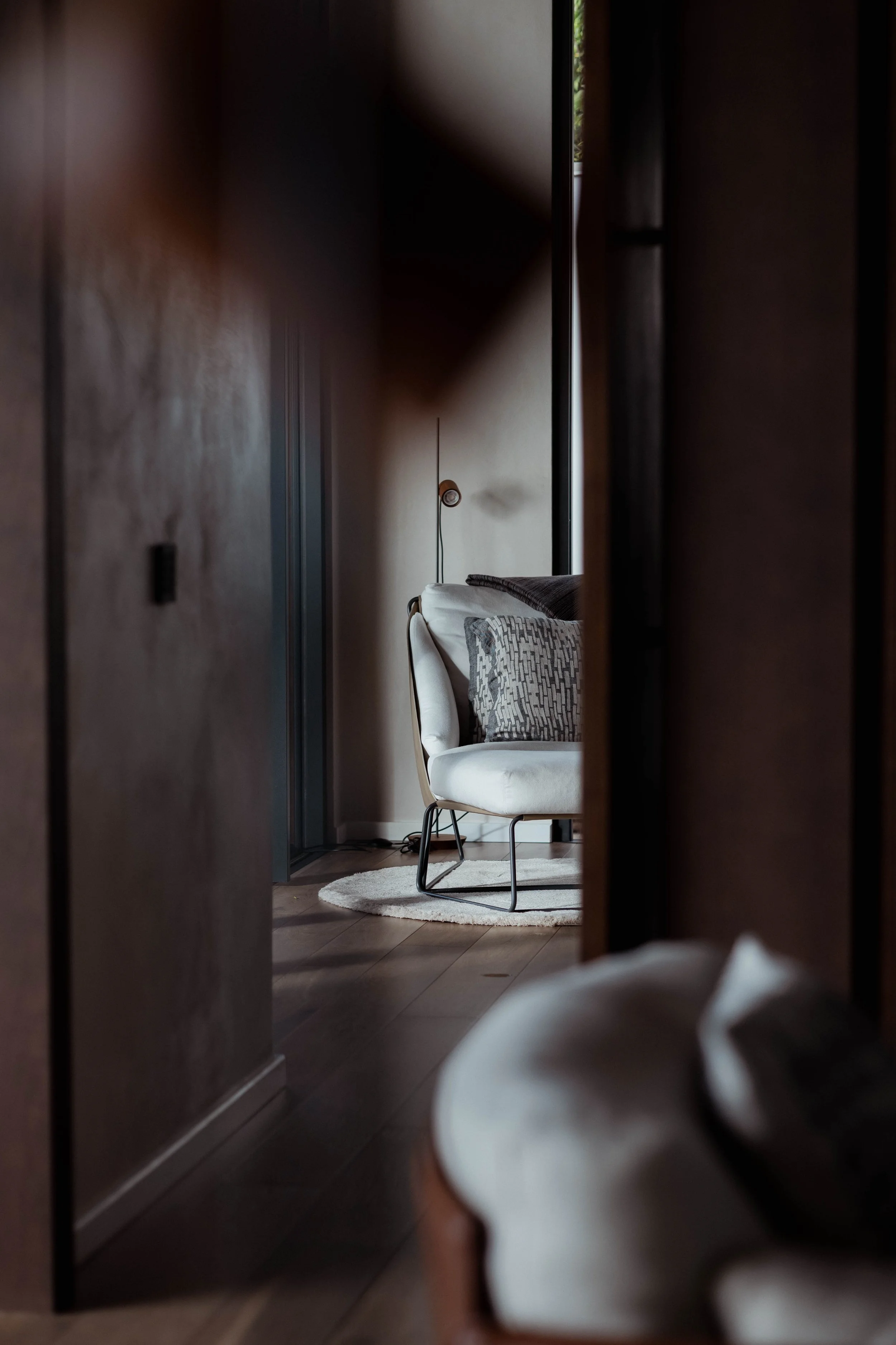 Vue d'un coin salon à travers une ouverture d'une porte, avec un fauteuil blanc, des coussins gris, un tapis rond blanc, et un mur avec une lampe déco