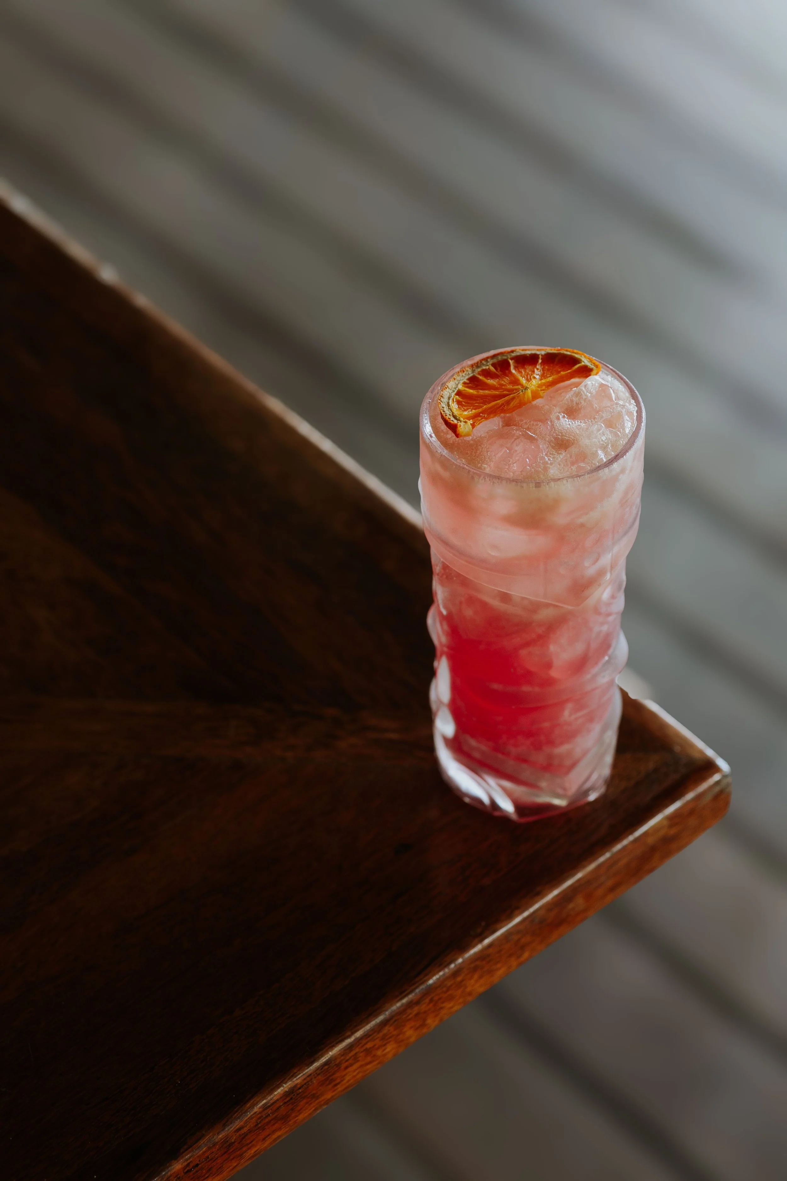 Un verre long avec une boisson rose, décorée d'une tranche de citron orange, posé sur une table en bois.