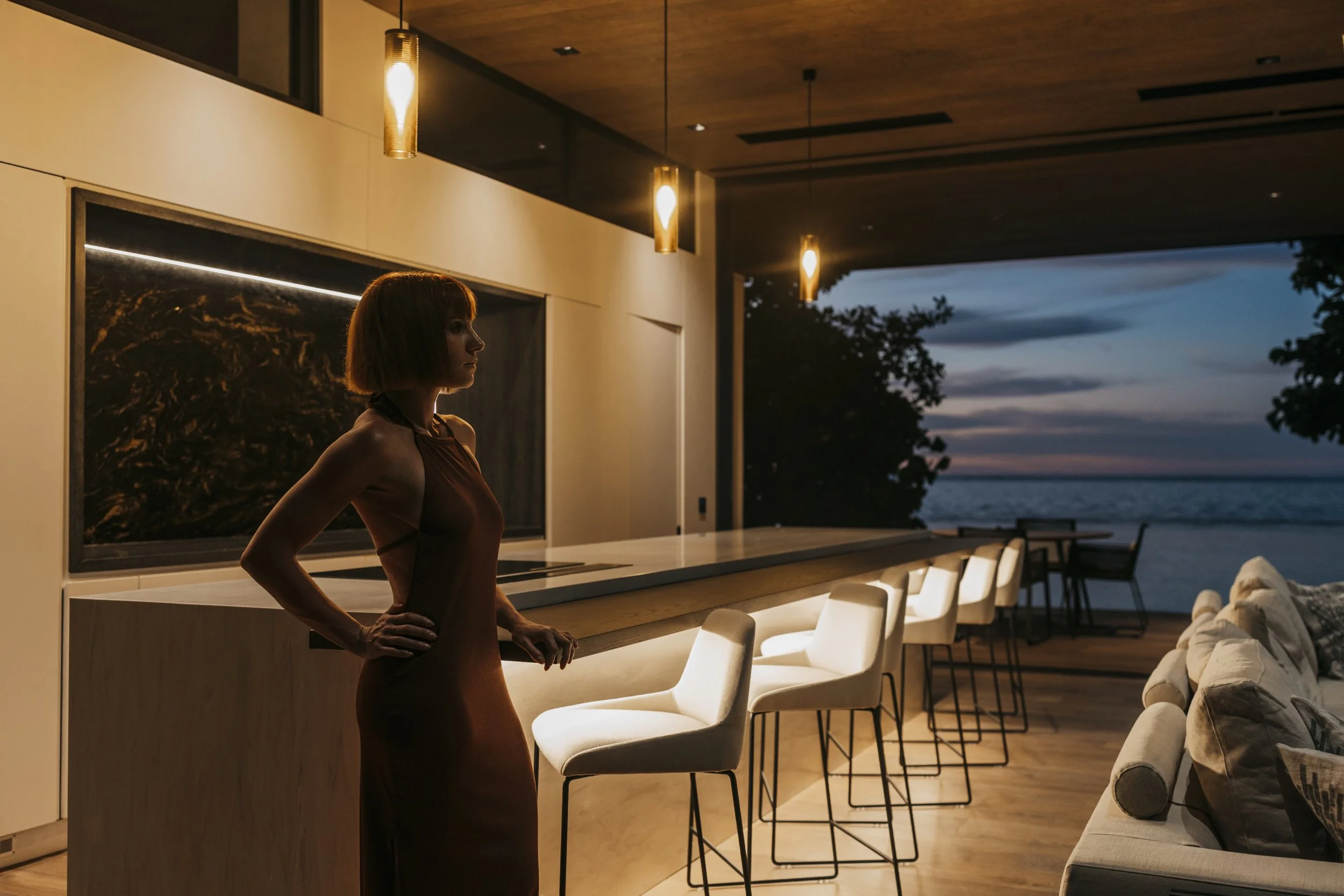 Une femme en robe marron se tient à l'intérieur d'une maison moderne au coucher du soleil, avec vue sur la plage et la mer extérieure, dans une pièce éclairée par des lampes suspendues.