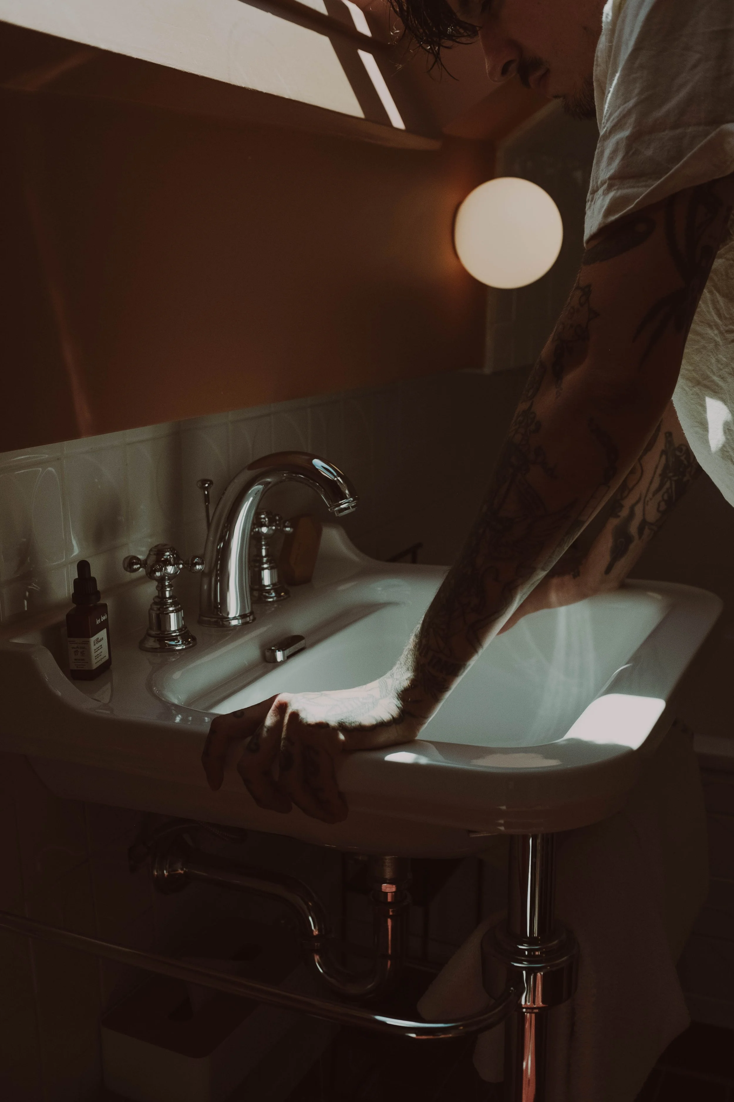 Une personne tatouée se penche sur un lavabo blanc dans une salle de bain, éclairée par une lumière chaude