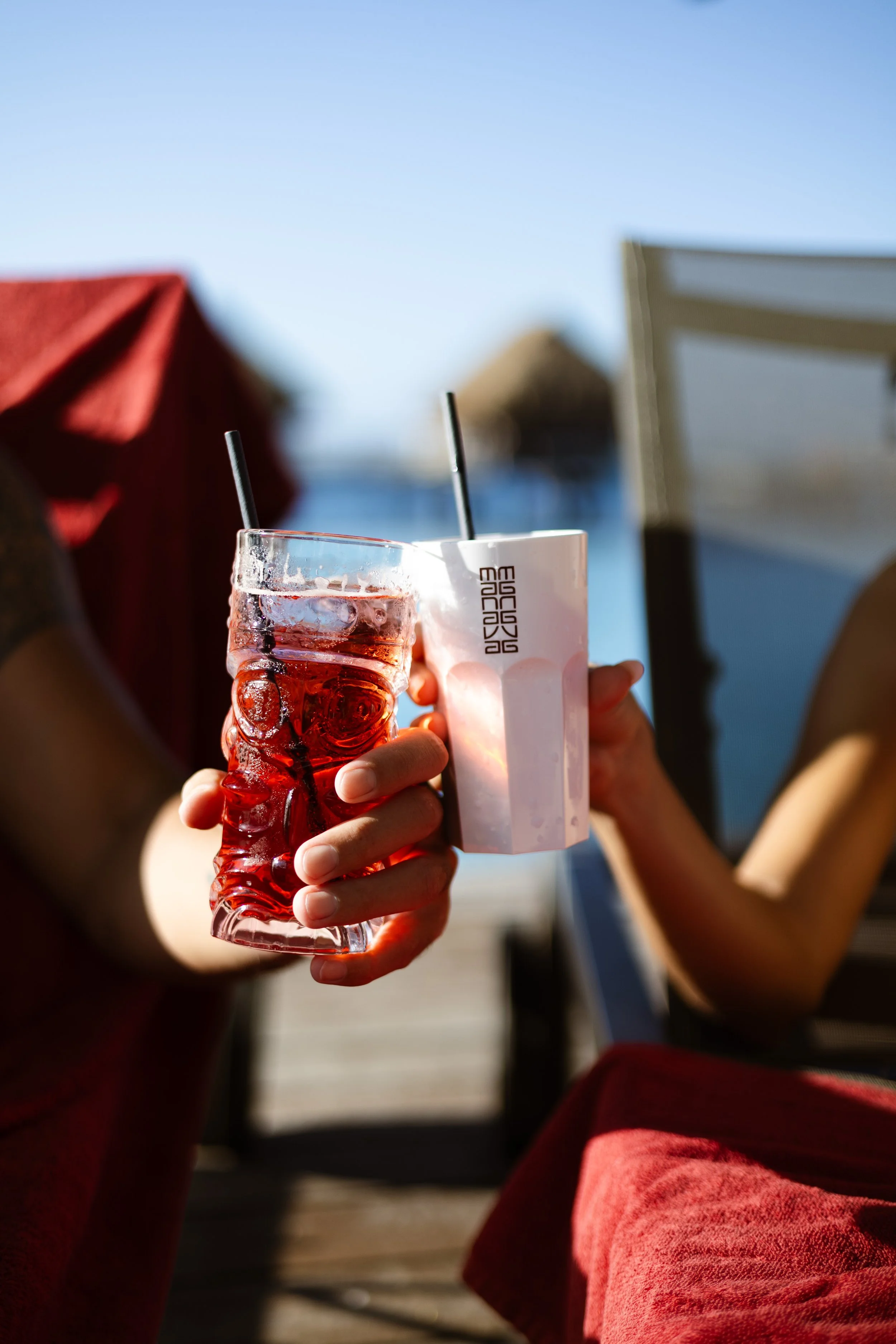 Deux personnes tiennent des verres à cocktail, l'un rouge avec des glaçons et l'autre blanc, en trinquant au bord d'une piscine avec la mer en arrière-plan, par une journée ensoleillée.