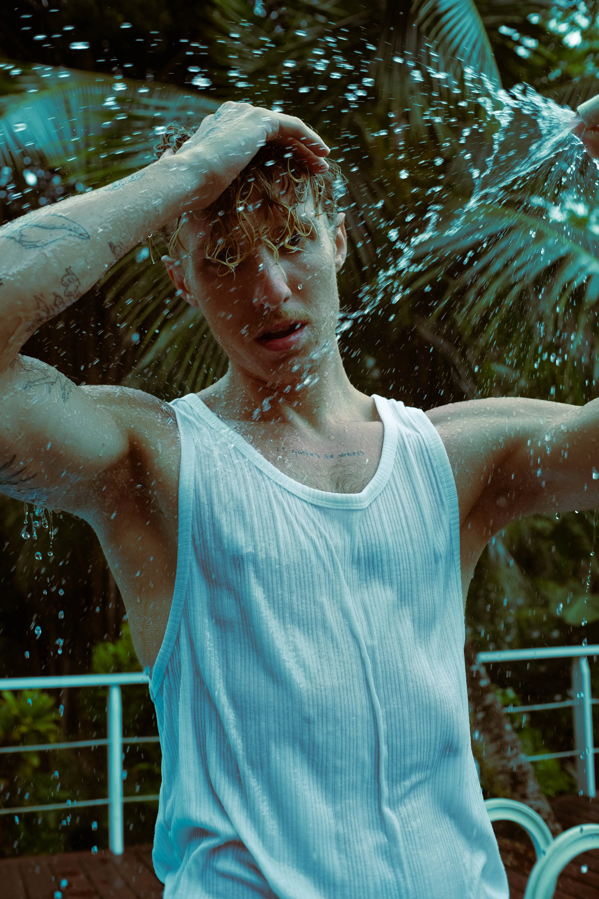 Jeune homme avec des cheveux bouclés mouillés, portant un débardeur blanc, sous une douche extérieure, avec des feuilles de palmier en arrière-plan.
