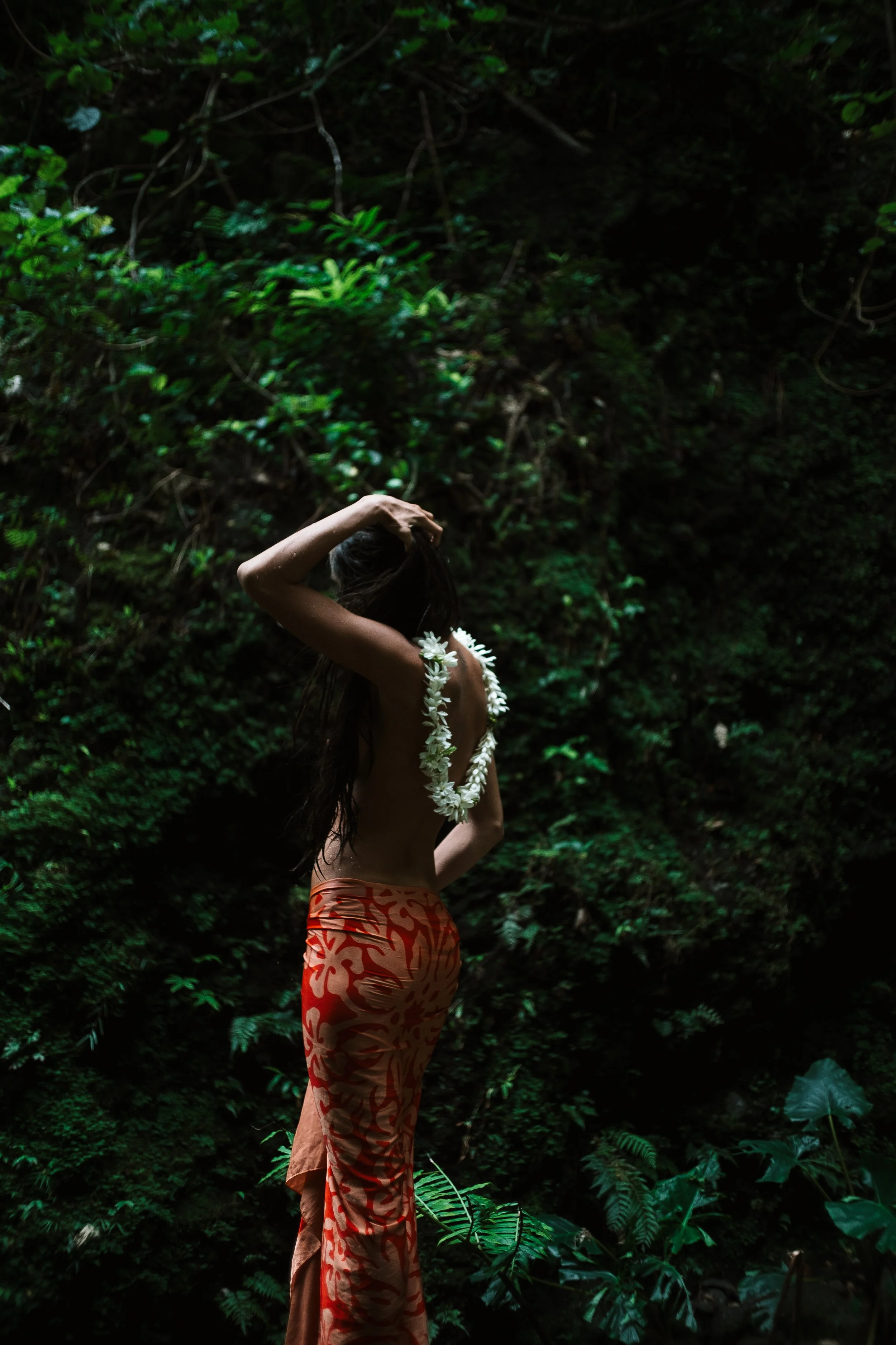 Une femme nue porte une couronne de fleurs et un sarong orange avec un motif rouge dans une forêt dense et sombre.