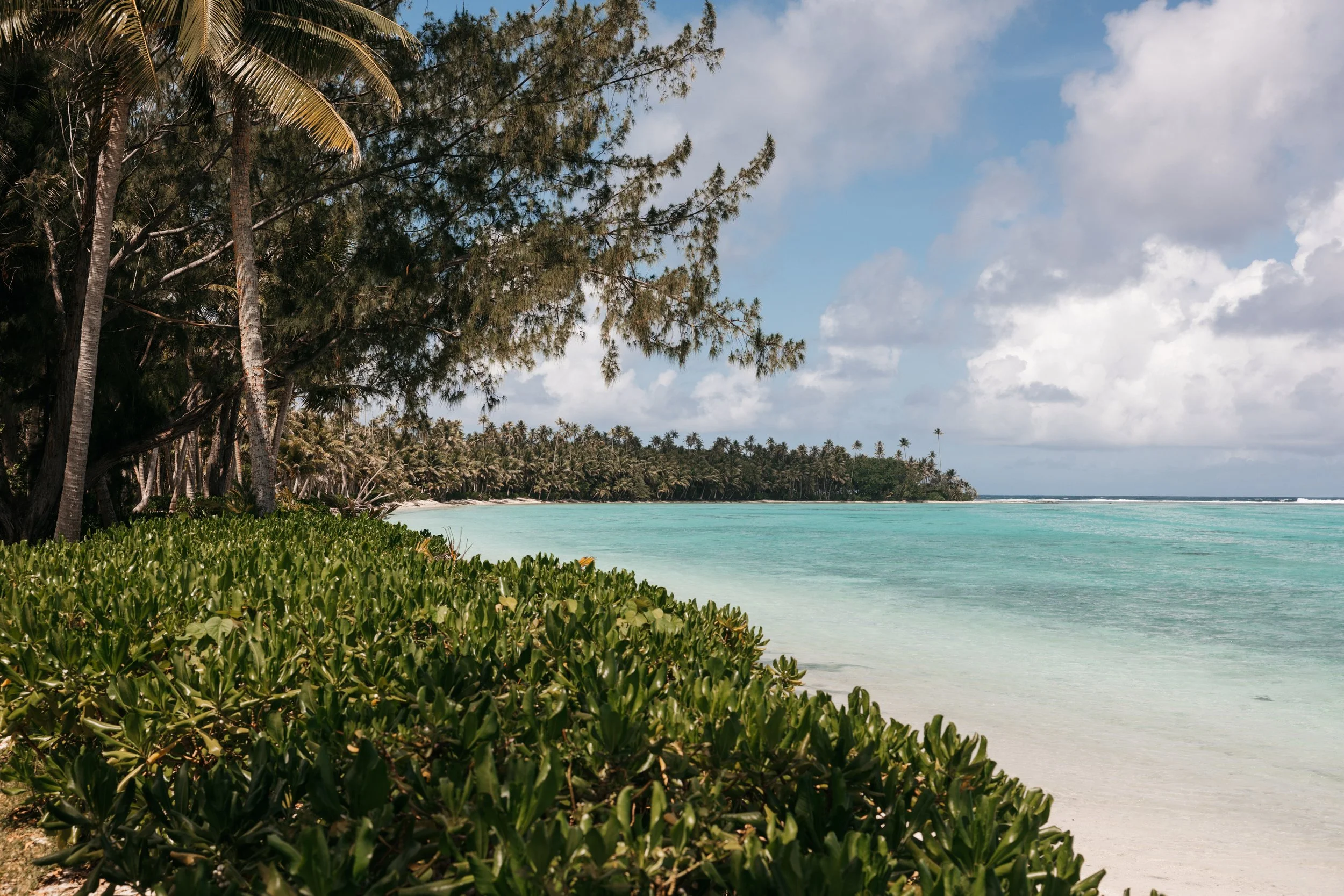 Plage de sable blanc avec une mer turquoise, des palmiers et un ciel partiellement nuageux.