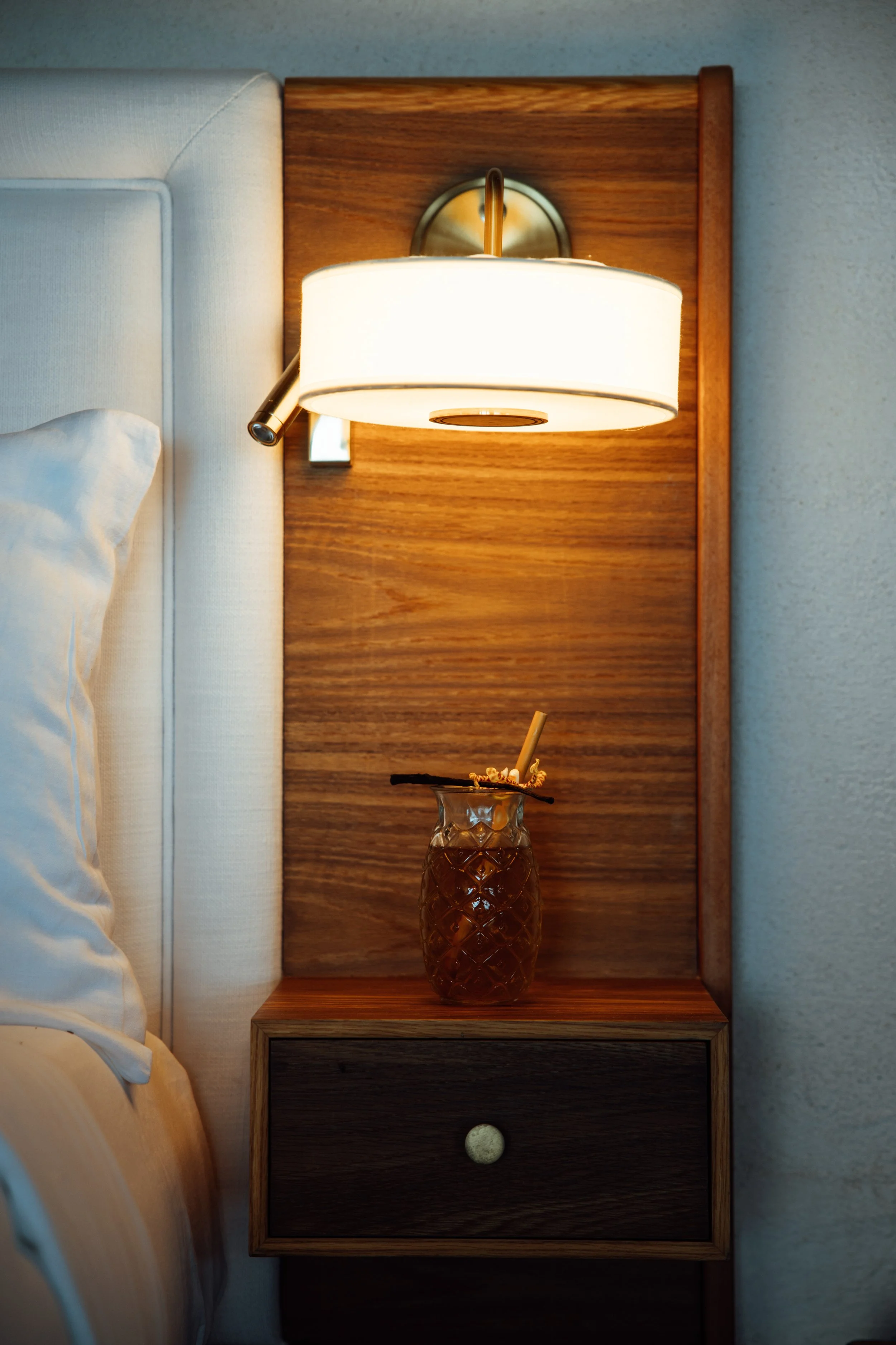 Lampe de chevet allumée, avec un vase en verre contenant une boisson et une paille, posé sur une table de nuit en bois.