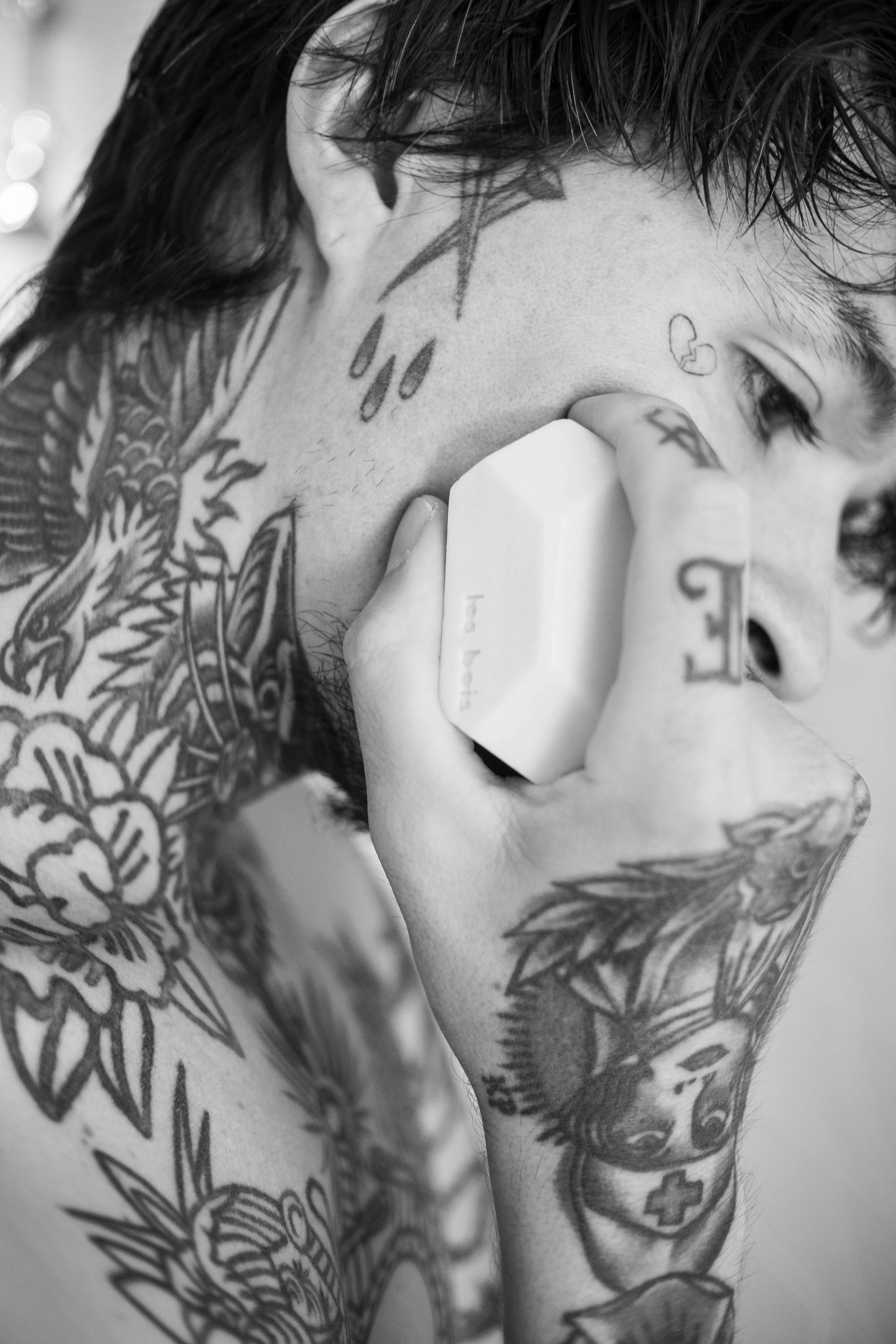 Un homme avec de nombreux tatouages parle au téléphone, tenant un téléphone blanc près de l'oreille, en noir et blanc.