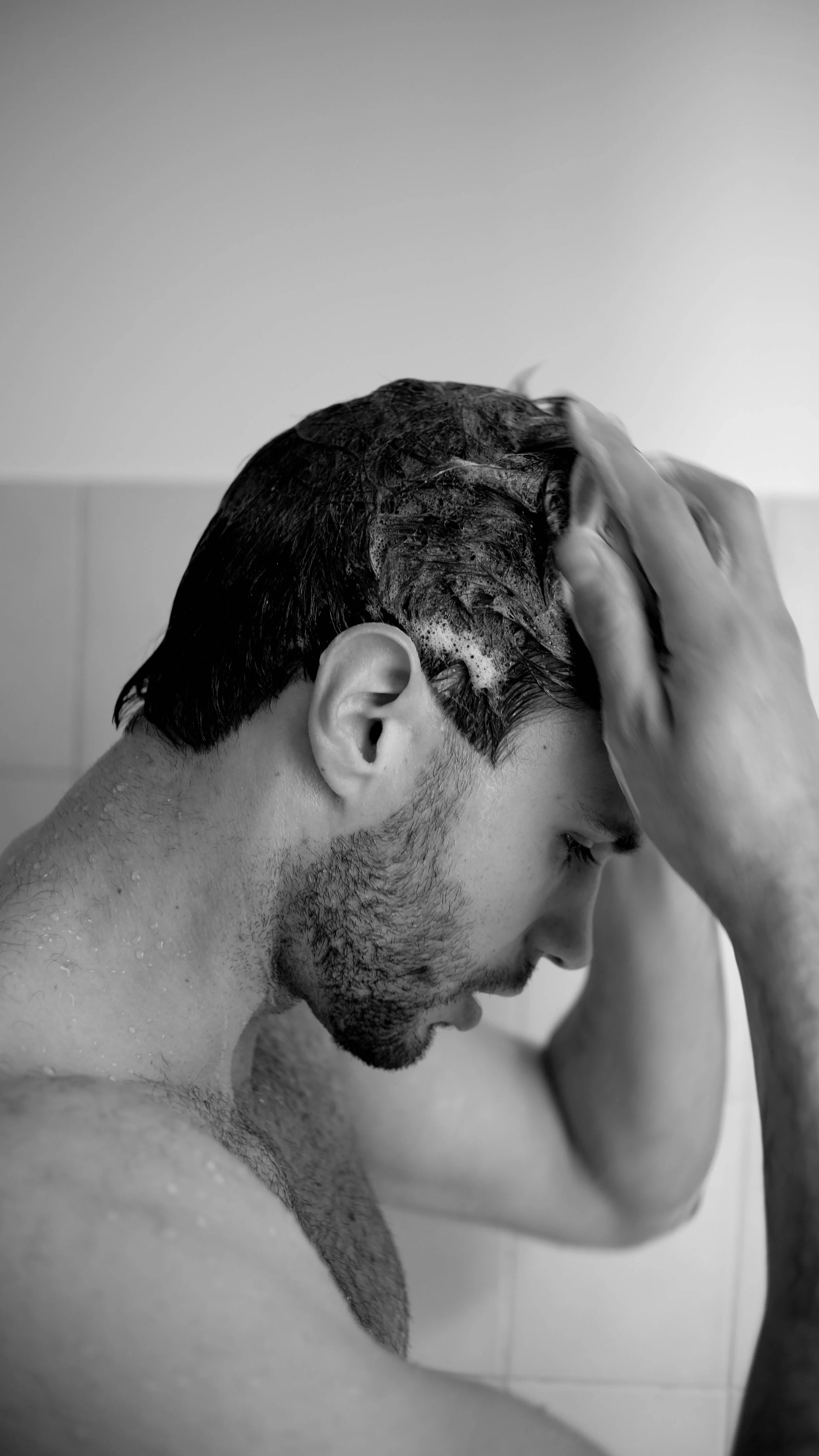 Un homme se rince les cheveux sous la douche, avec de la mousse sur la tête, en noir et blanc.