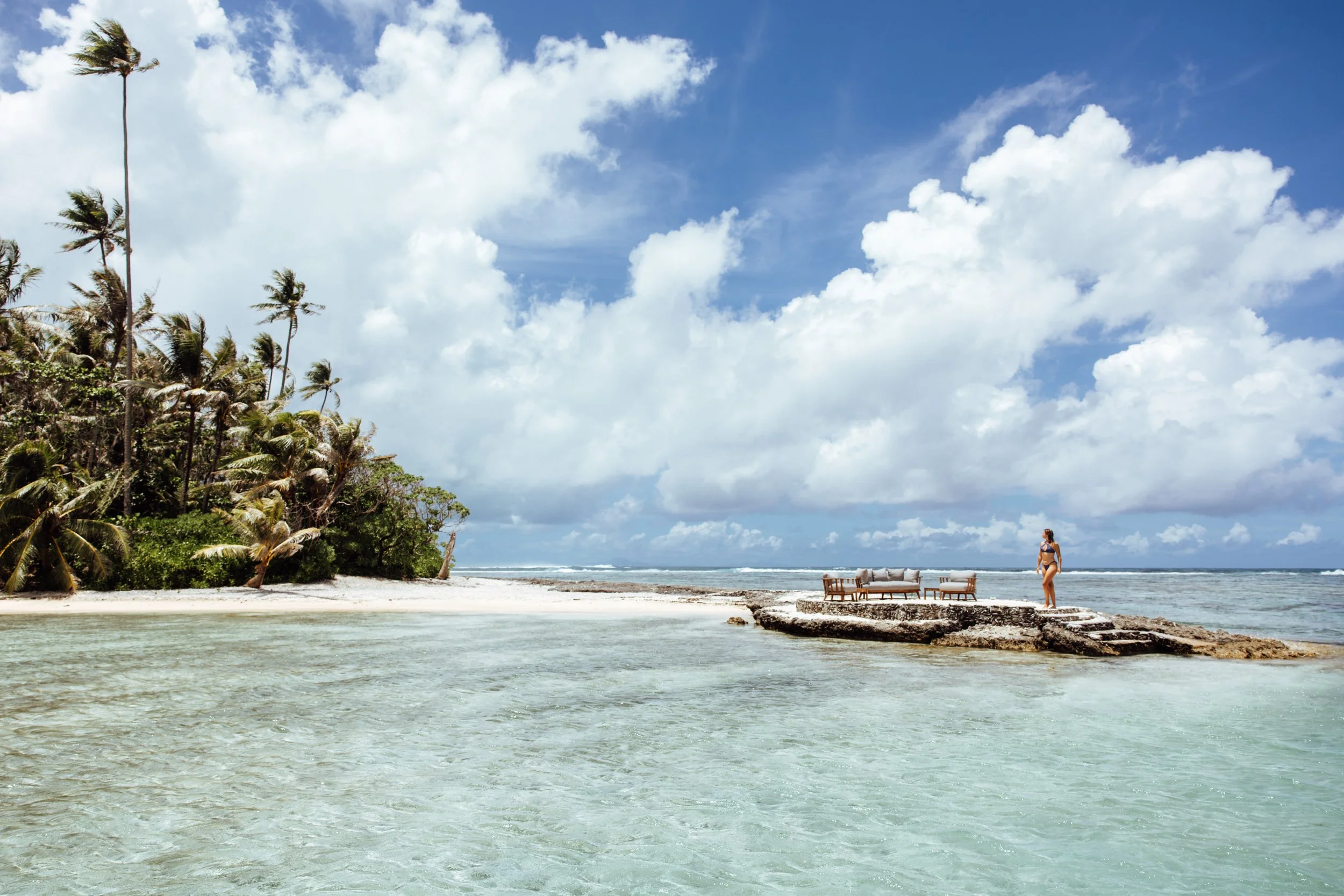 Plage tropicale avec sable blanc, eau claire, palmiers, ciel nuageux, un transat, un canapé, une femme en maillot de bain.