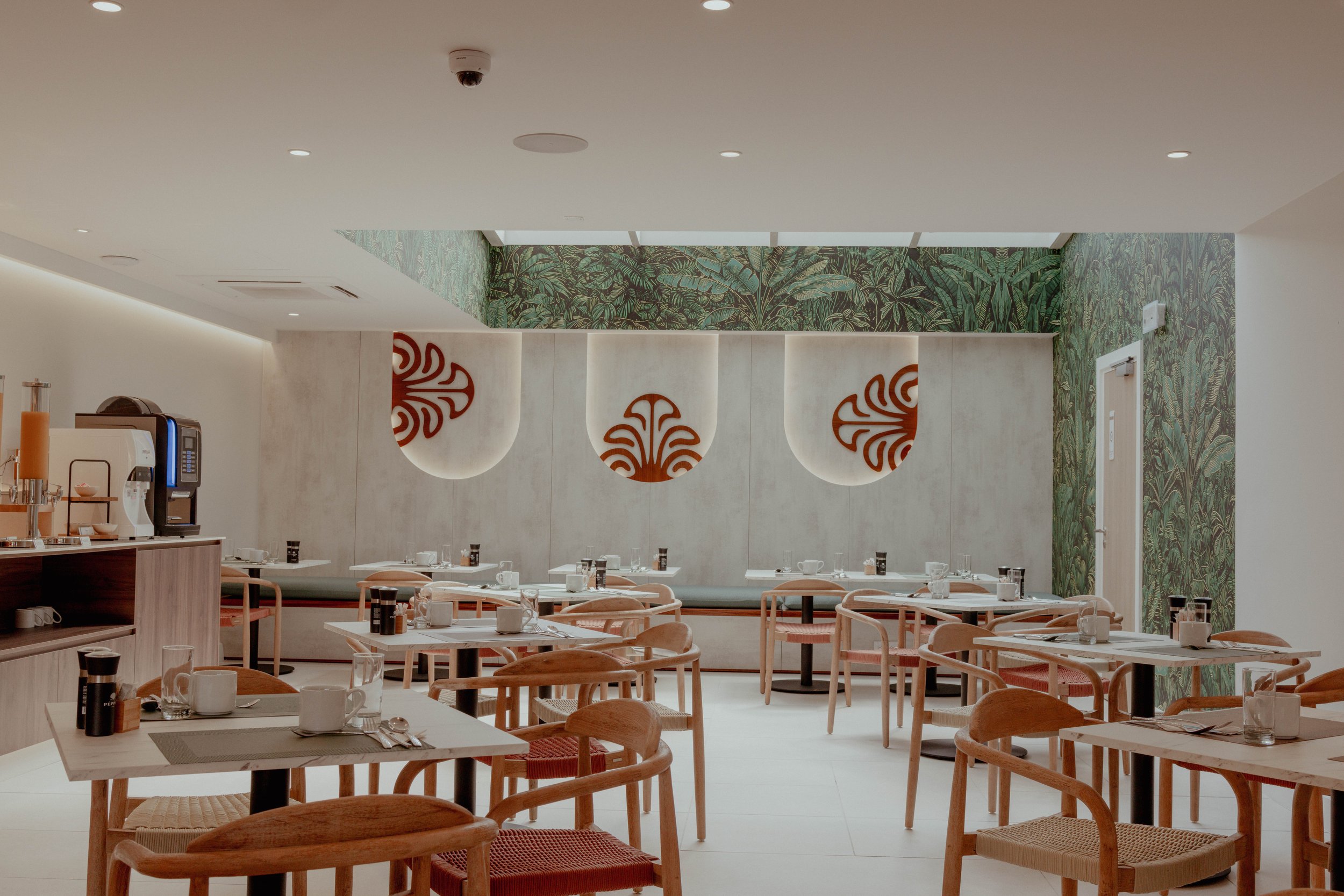 Salle à manger d'un restaurant moderne avec tables, chaises en bois, décoration verte et motifs rouges sur la muraille beige.
