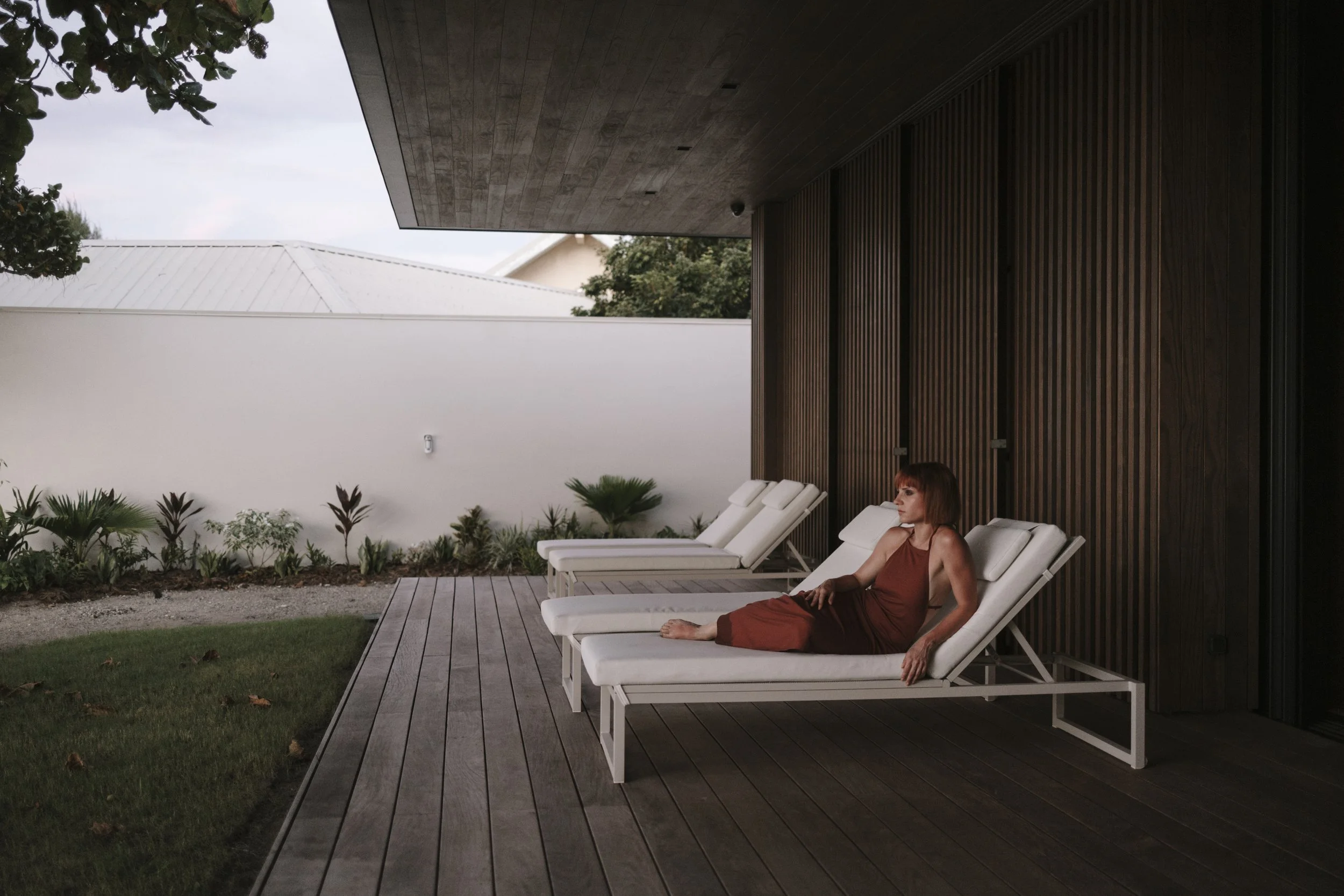Une femme assise sur un bain de soleil blanc sur une terrasse en bois, avec un mur en bois foncé derrière elle et un jardin avec des plantes à l'arrière-plan.
